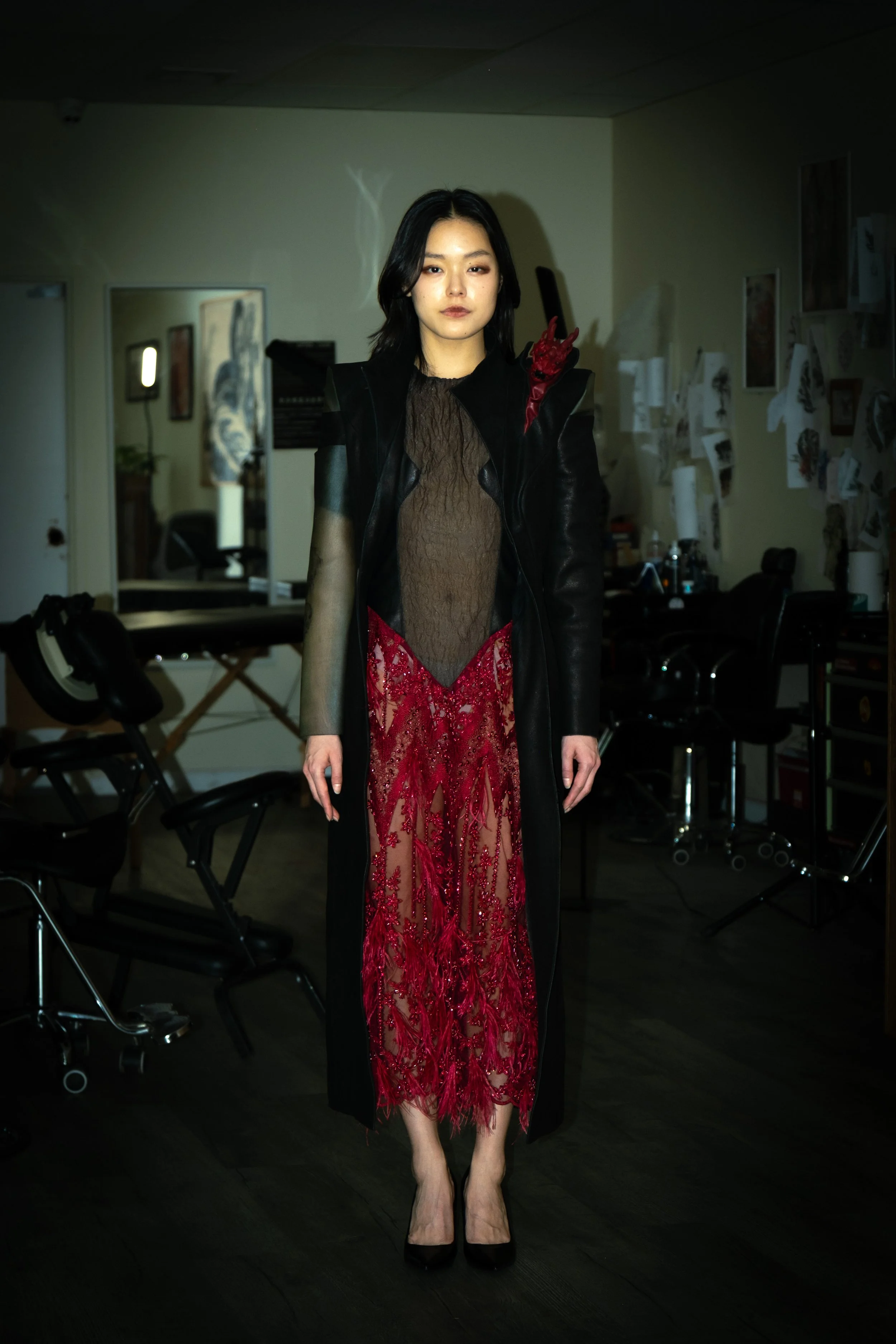 A woman with black hair and makeup wearing a black jacket with shoulder details, a sheer lace brown top, and a red lace dress with feather details, standing in a room with art on the walls and various furniture.