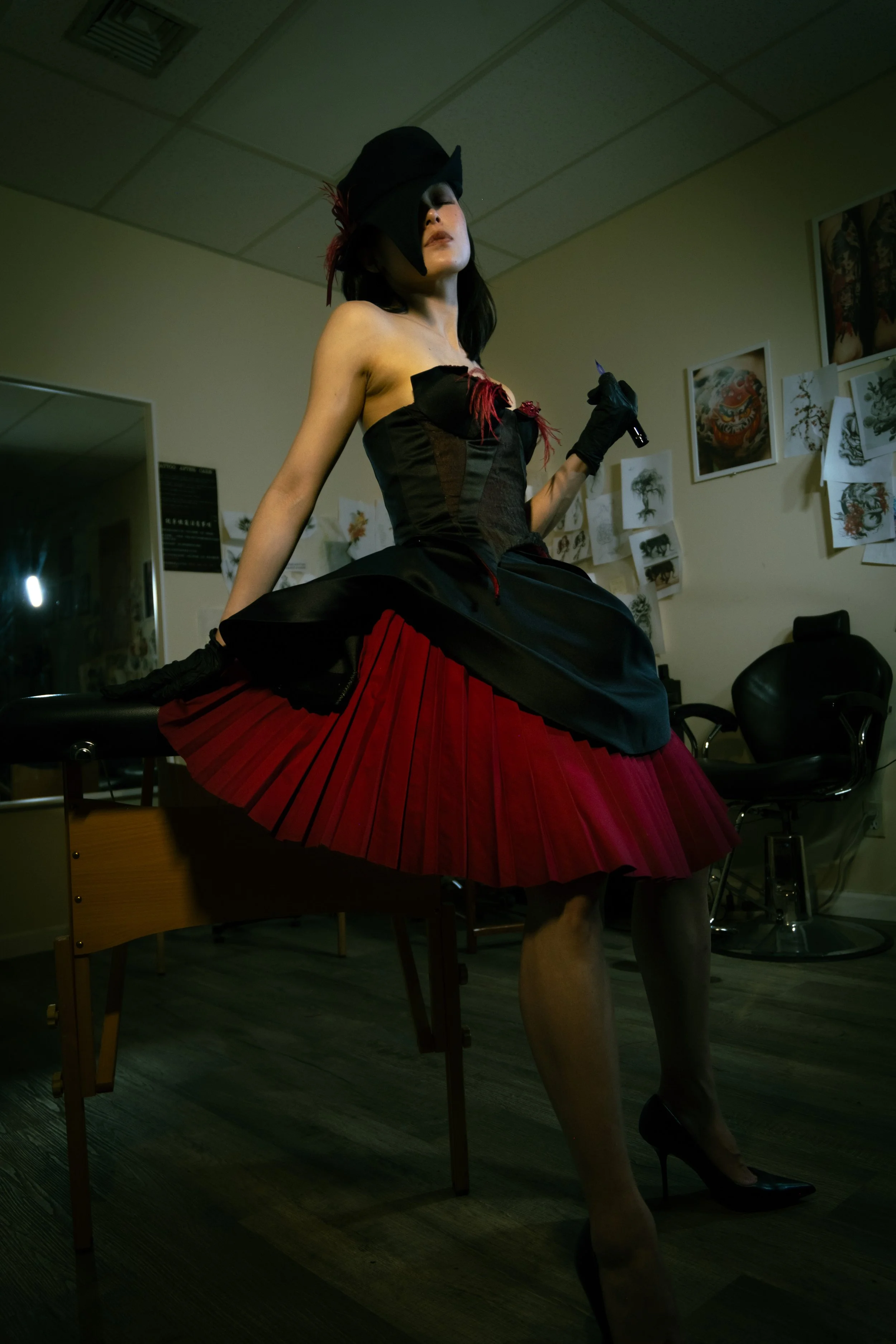 A woman dressed in a dark corset, red pleated skirt, and high heels, wearing a black hat with a feather, holding a tattoo machine, and sitting on a table in an art studio with tattoo designs on the wall.