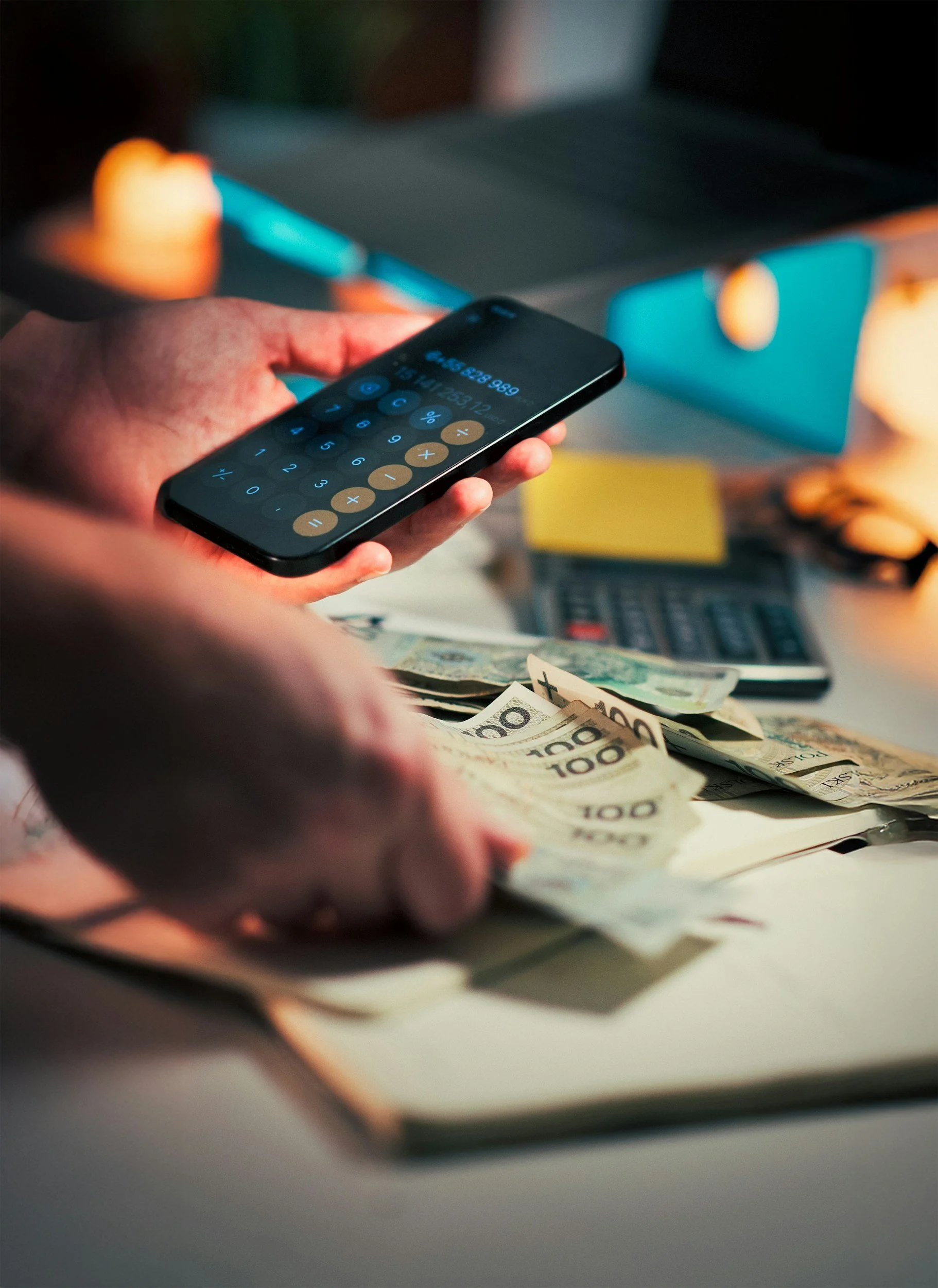 A person holding a smartphone displaying a calculator app, with US dollar bills and a calculator on a desk in the background.
