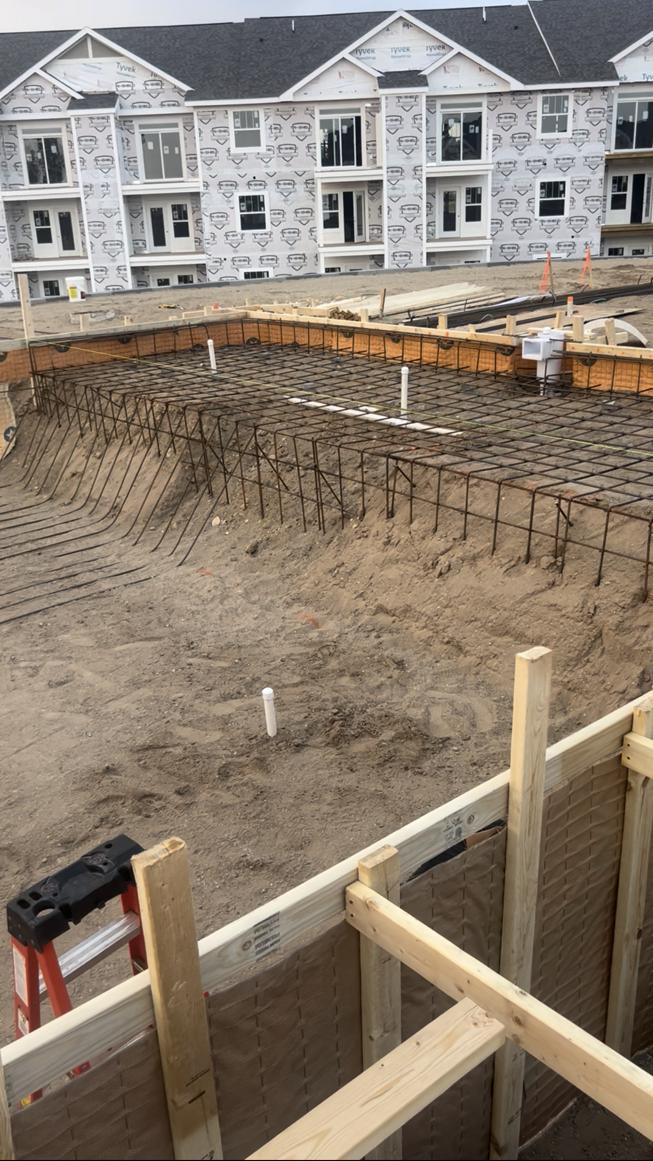 Construction site with steel rebar forming the foundation for a building's concrete slab, surrounded by dirt and wooden framing, with an unfinished multi-story building in the background.