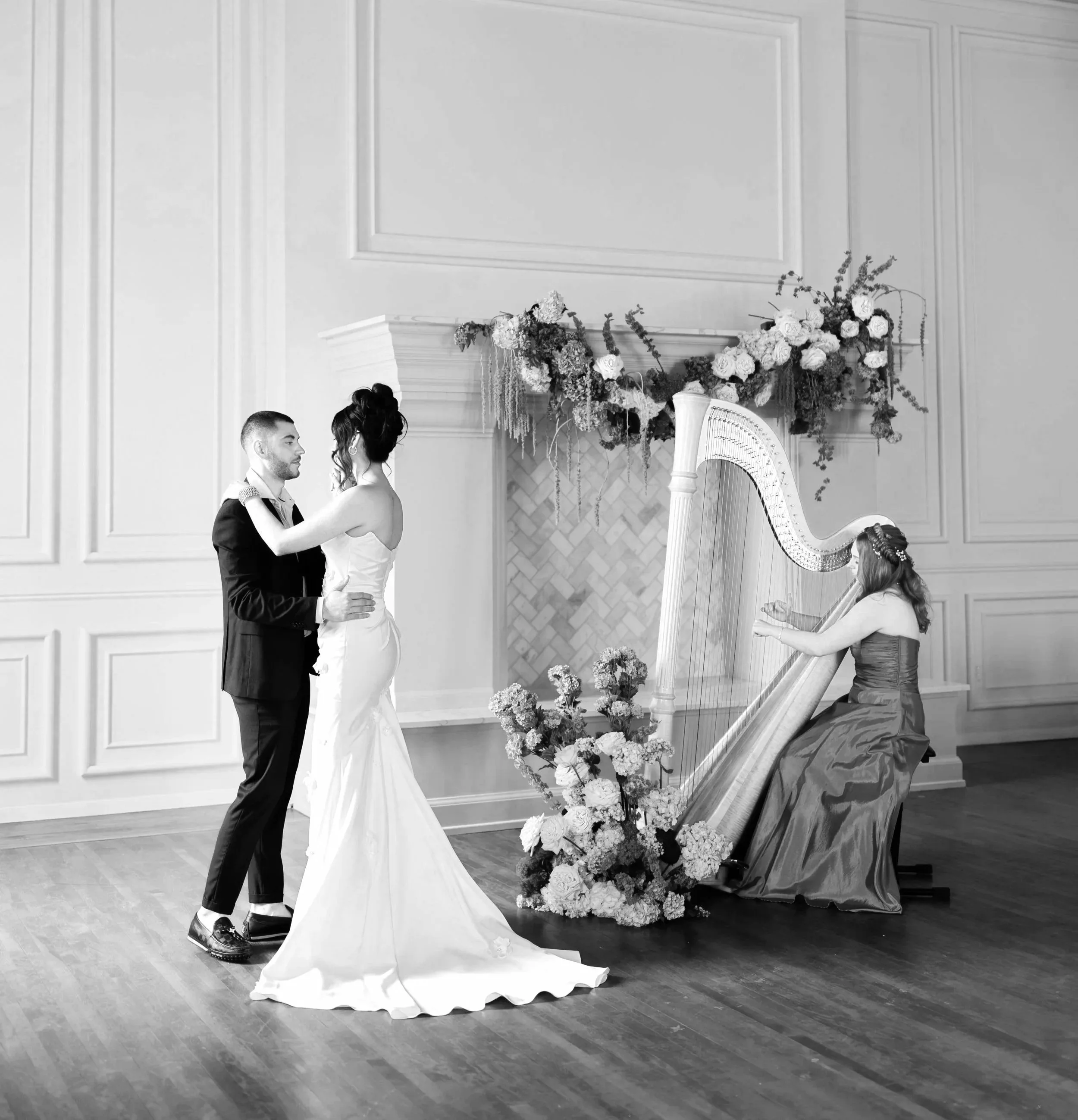 A couple dressed in formal attire dancing together in a room decorated with floral arrangements and a harp, with a woman in a gown playing the harp.