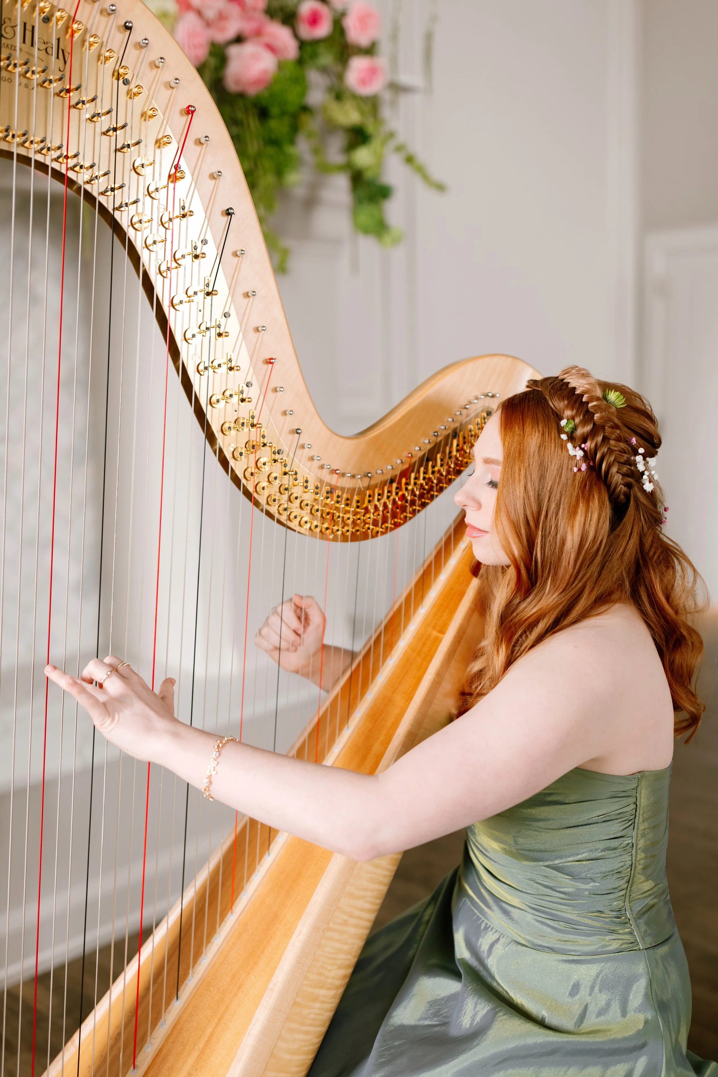 A woman with long red hair styled with flowers, wearing a green satin dress, playing a harp in a bright room with pink flowers in the background.