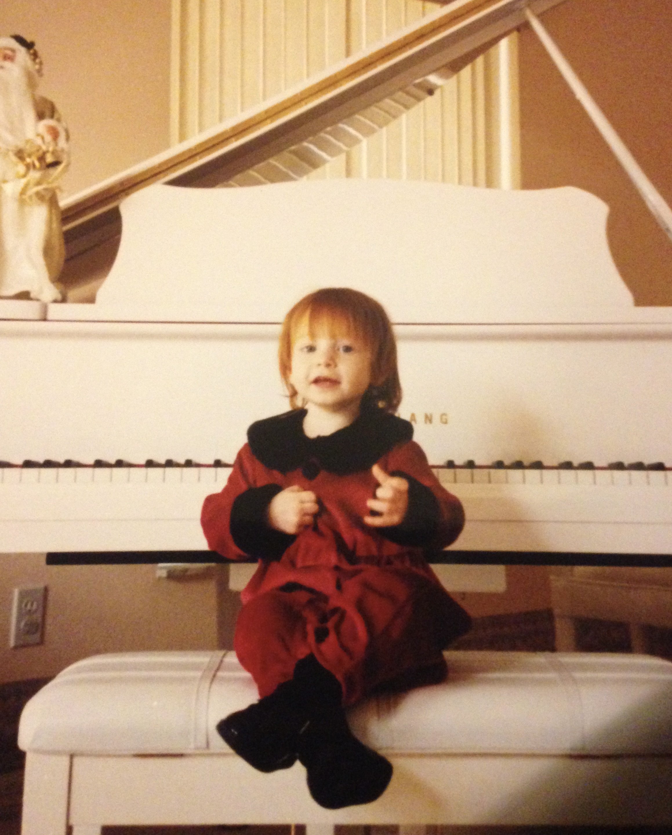 A young boy with red hair and a black winter hat is sitting on a bed in a bedroom, in front of a white piano. The boy is wearing a red jacket with black accents and has a curious expression. The room has warm-colored walls, and the piano has a decorative open sheet music stand.