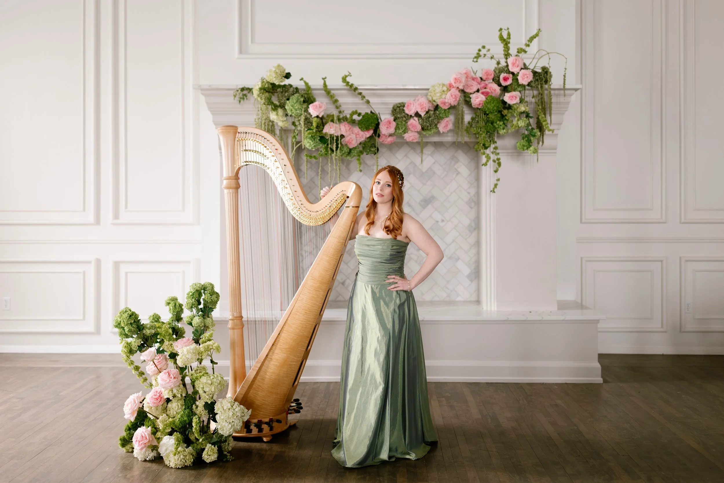 A woman in a green dress standing beside a harp, with a floral arrangement of pink and white flowers in front of her and above her, in a white room with paneled walls.