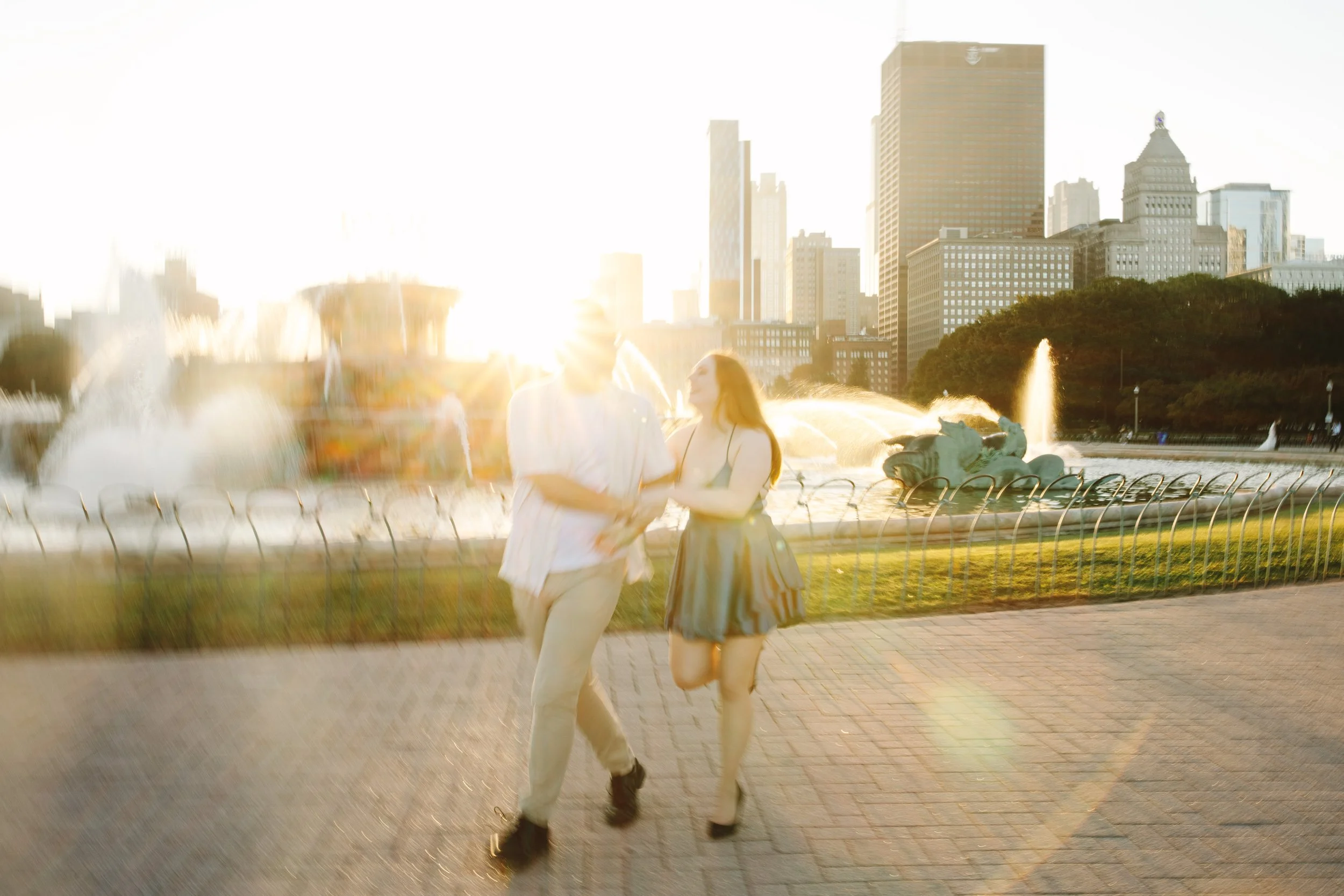 Two women walking in a city park during sunset with fountains and high-rise buildings in the background.