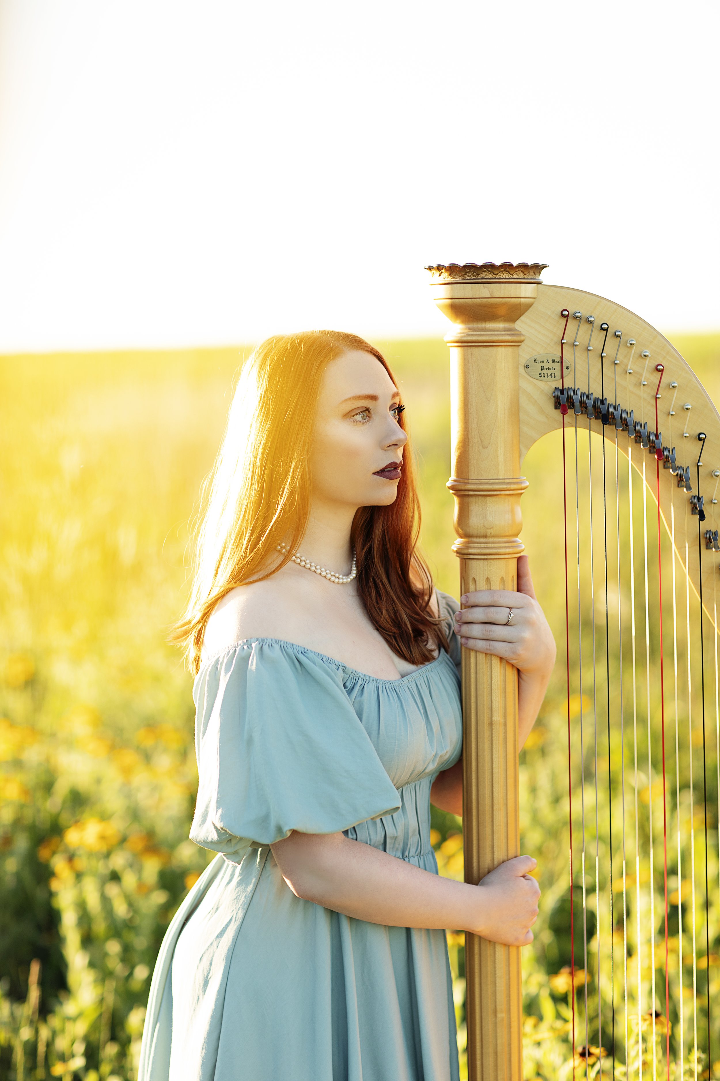 A woman with long red hair wearing a light blue off-shoulder dress and pearl necklace, standing in a field of yellow flowers, holding a large wooden harp with a tall column and a curved frame.