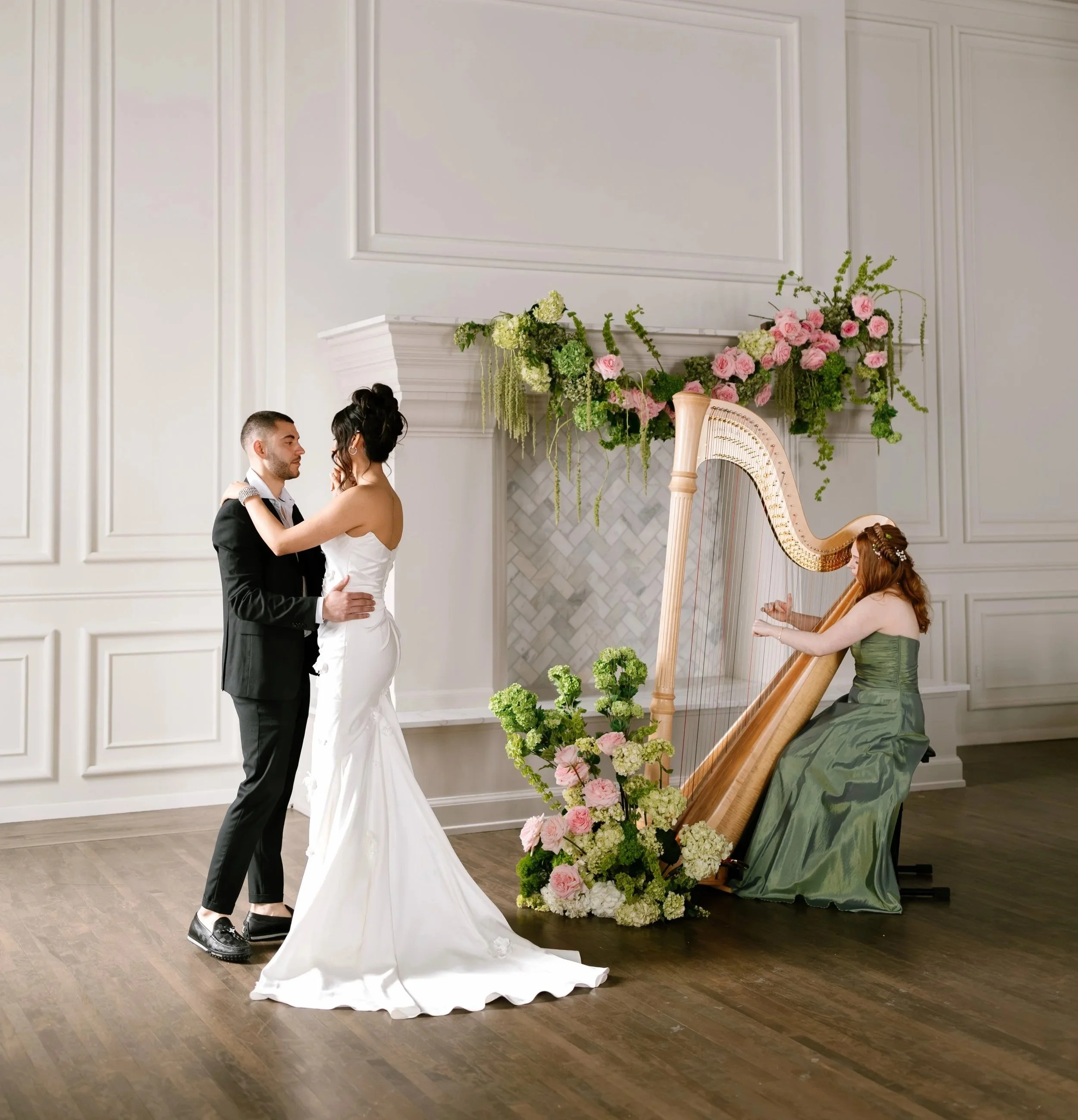 A couple is dancing during a wedding ceremony, with a harpist playing nearby. The ceremony takes place in an elegant room with white paneled walls and a marble fireplace decorated with pink and white flowers and greenery.