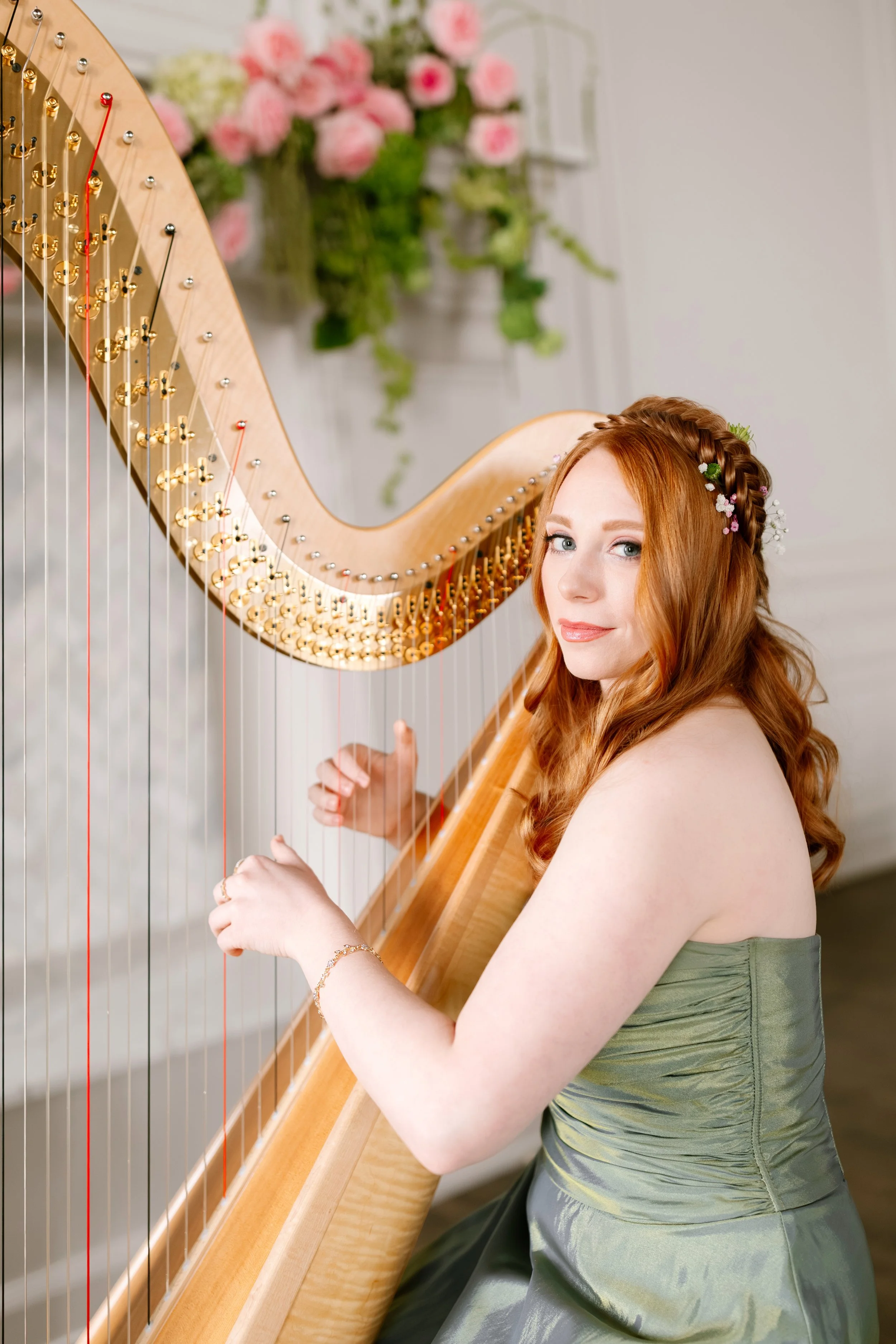 Red-haired woman in a green strapless dress playing a wooden harp with pink flowers on her hair, standing indoors.