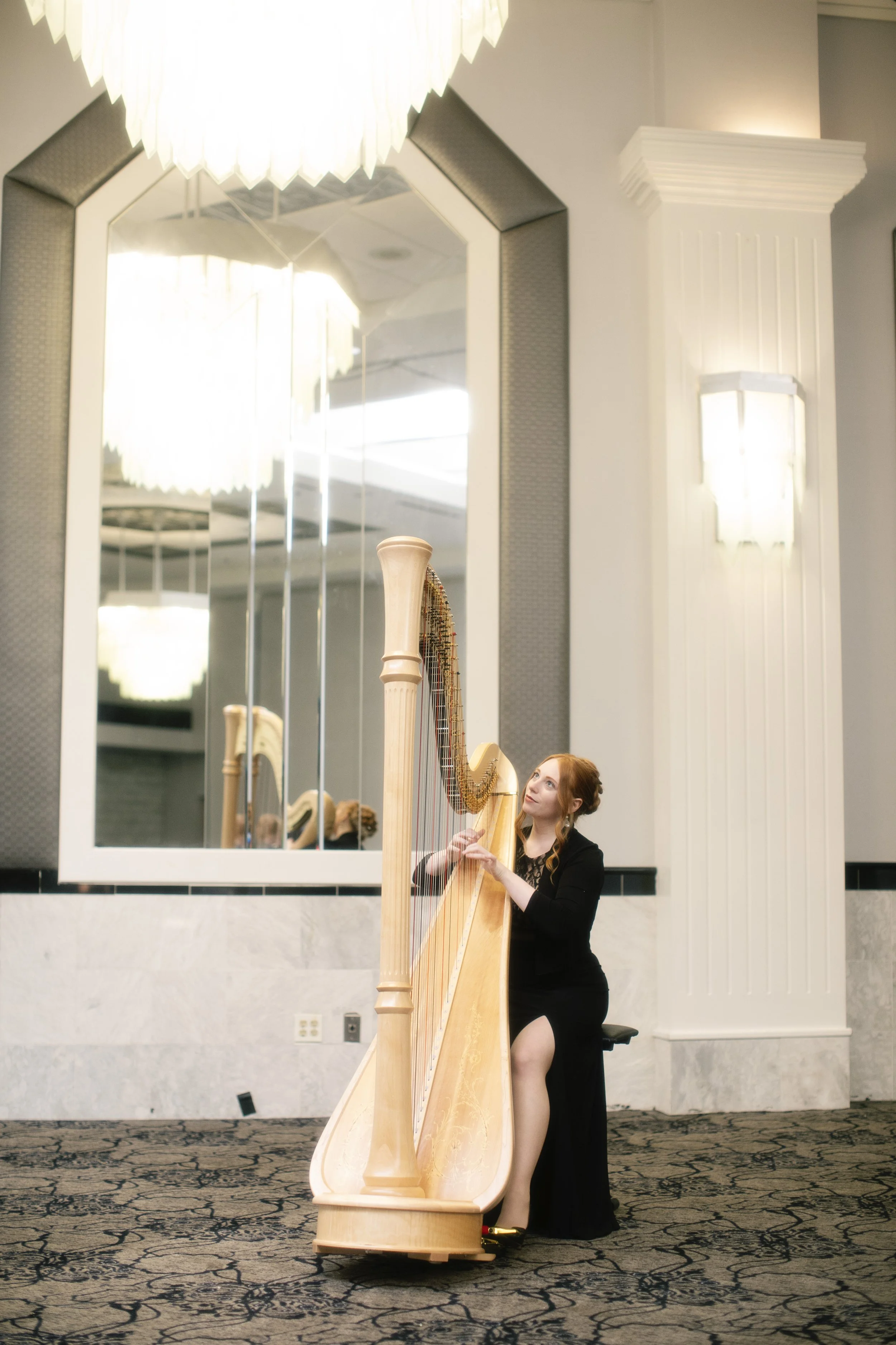 A woman in a long black dress playing a harp in a hotel lobby with a large mirror and decorative wall sconces.
