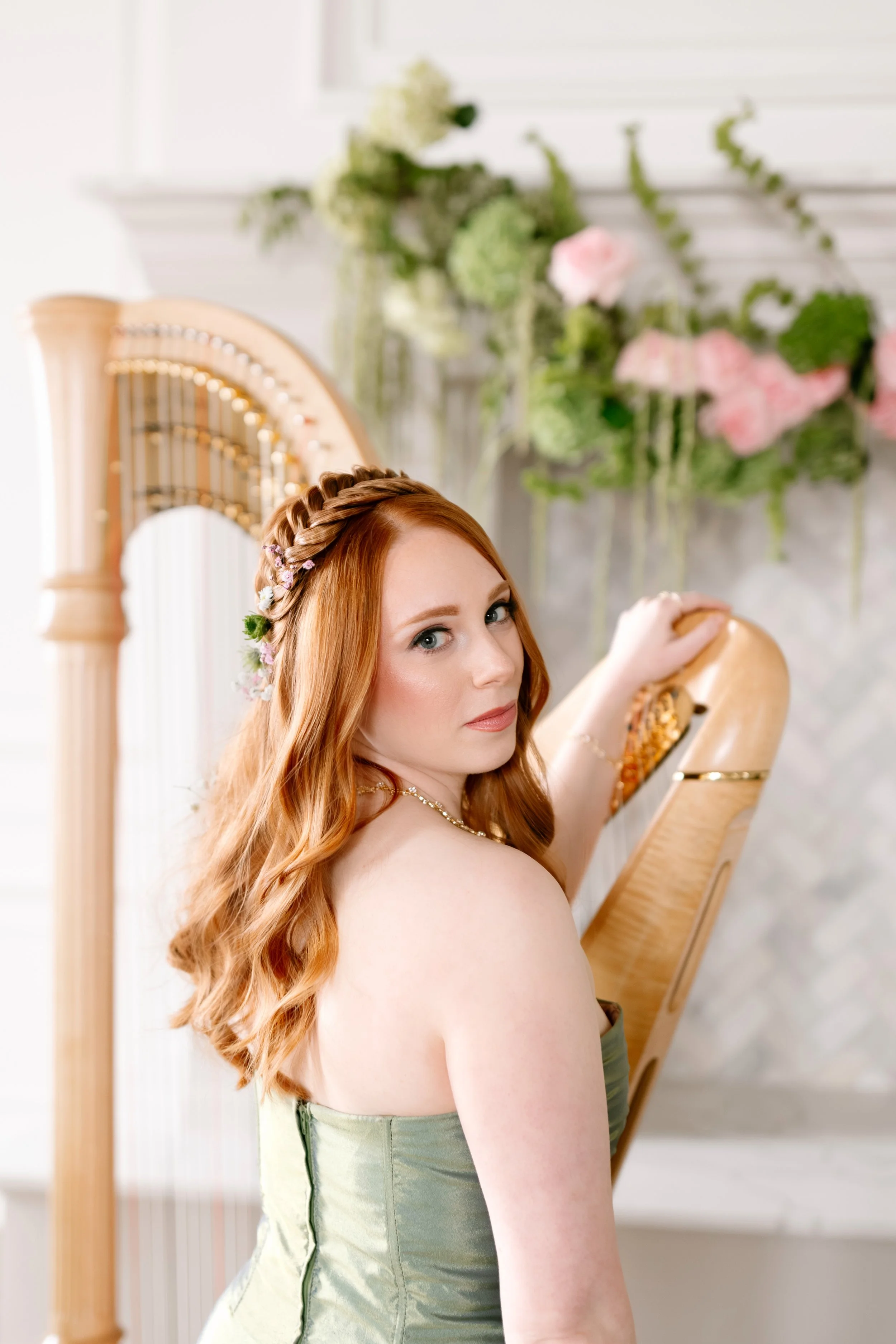 A woman with long red hair and a floral crown holds a harp, standing in front of a white wall with flowers.