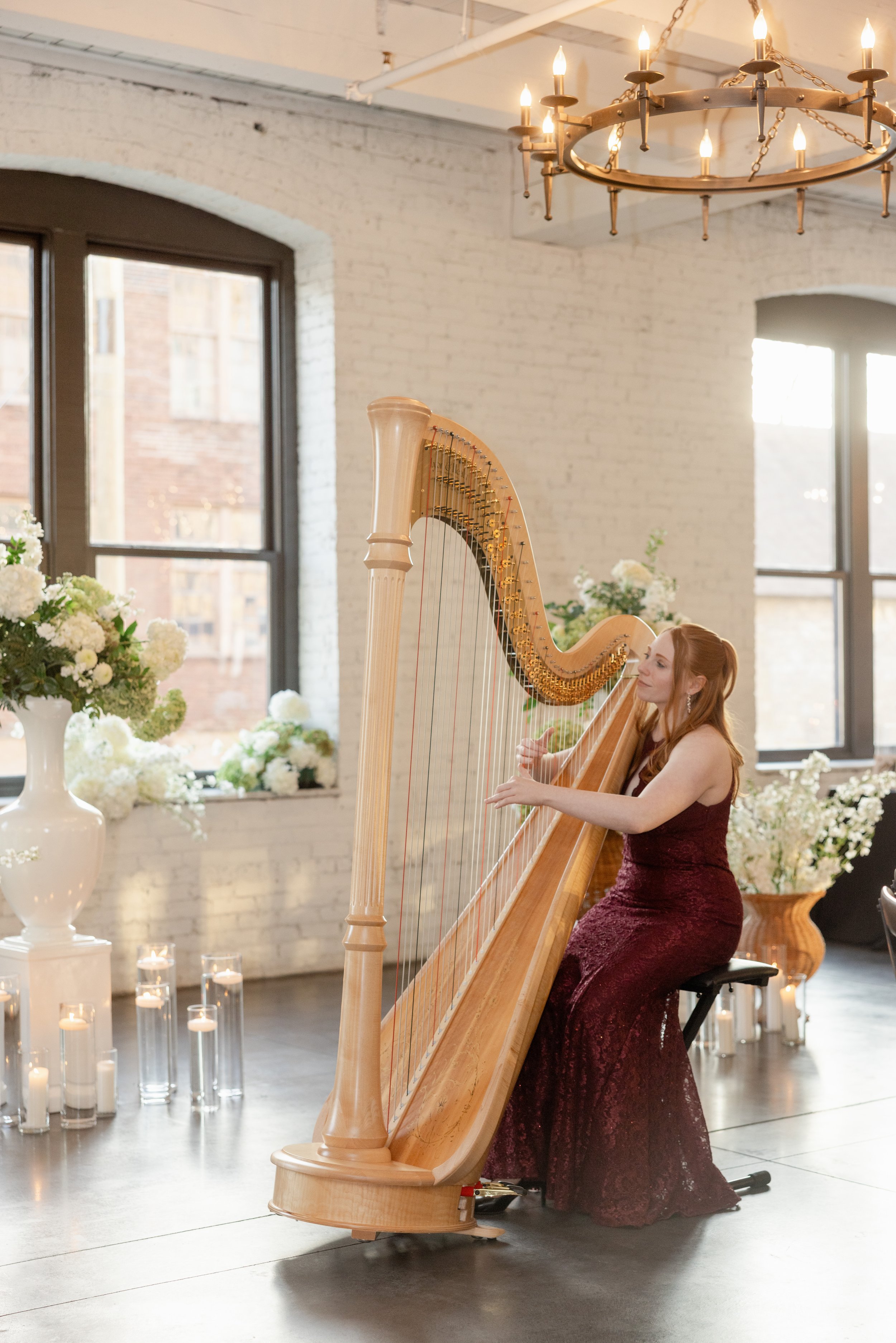 A woman in a burgundy gown playing a harp in a decorated venue with white brick walls, large windows, and a chandelier.