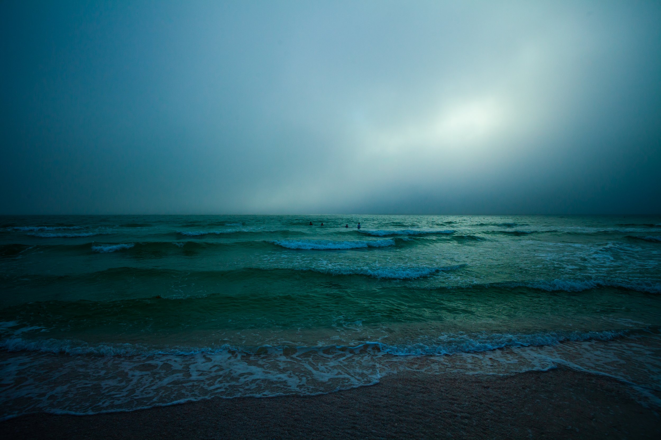 A view of the ocean under a cloudy sky with waves approaching the shore, and a few people swimming in the distance.