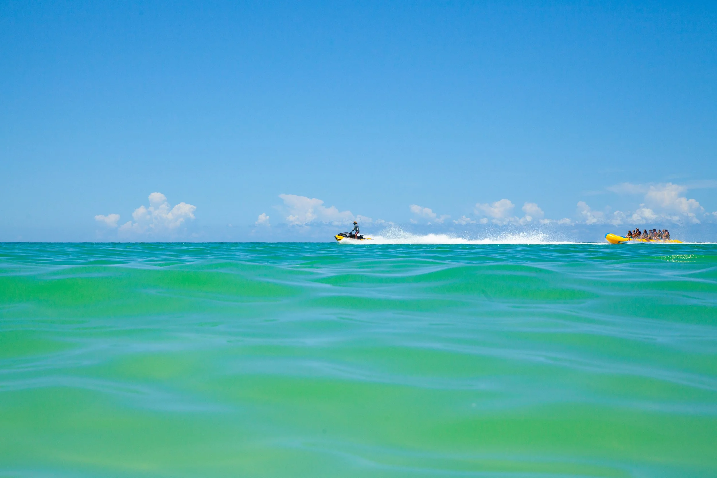 A person riding a jet ski and pulling a group of people on a yellow inflatable raft on a clear, sunny day at the ocean.