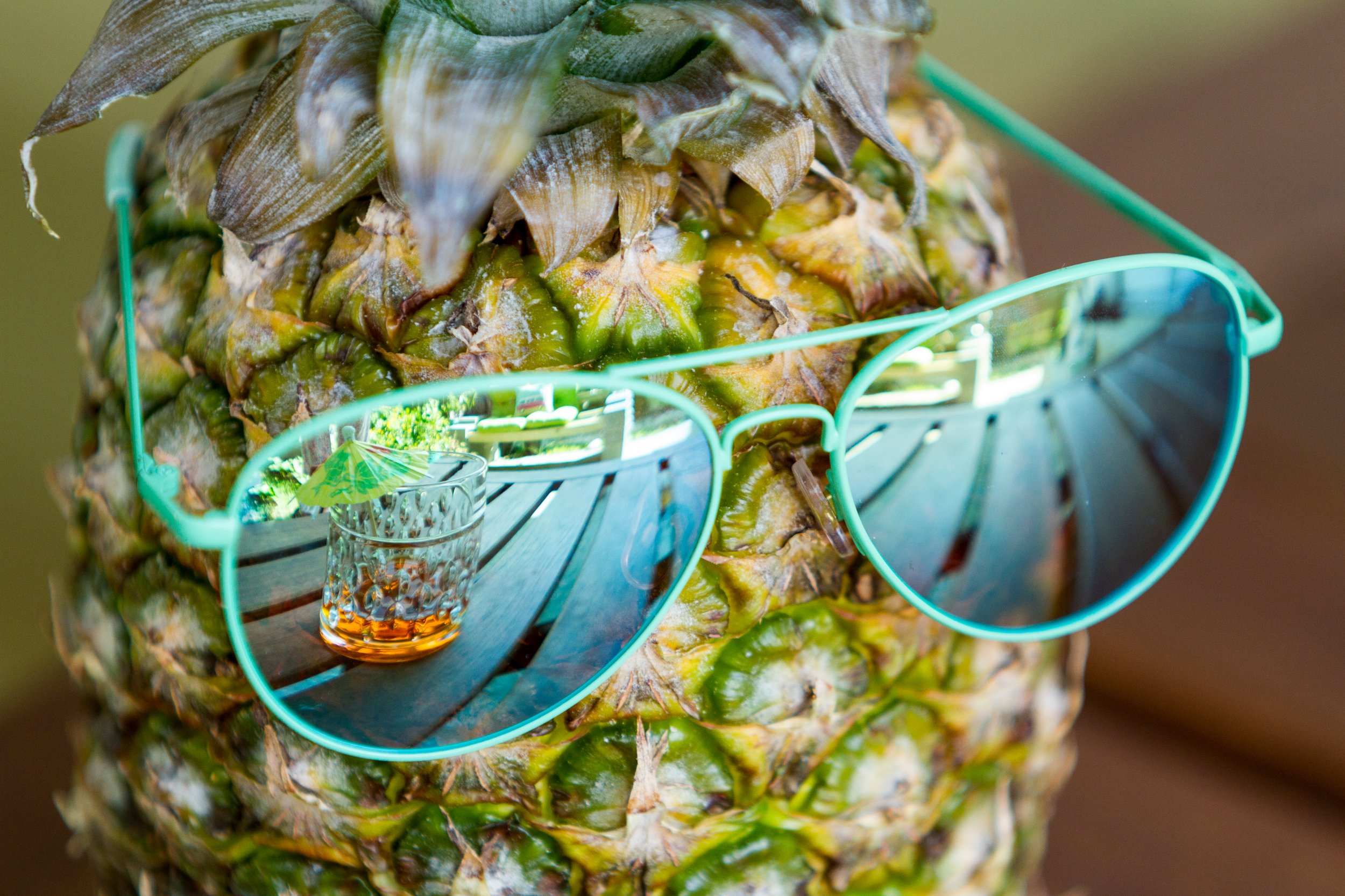 A pineapple wearing turquoise sunglasses with a reflection of a drink with an umbrella on the lenses.