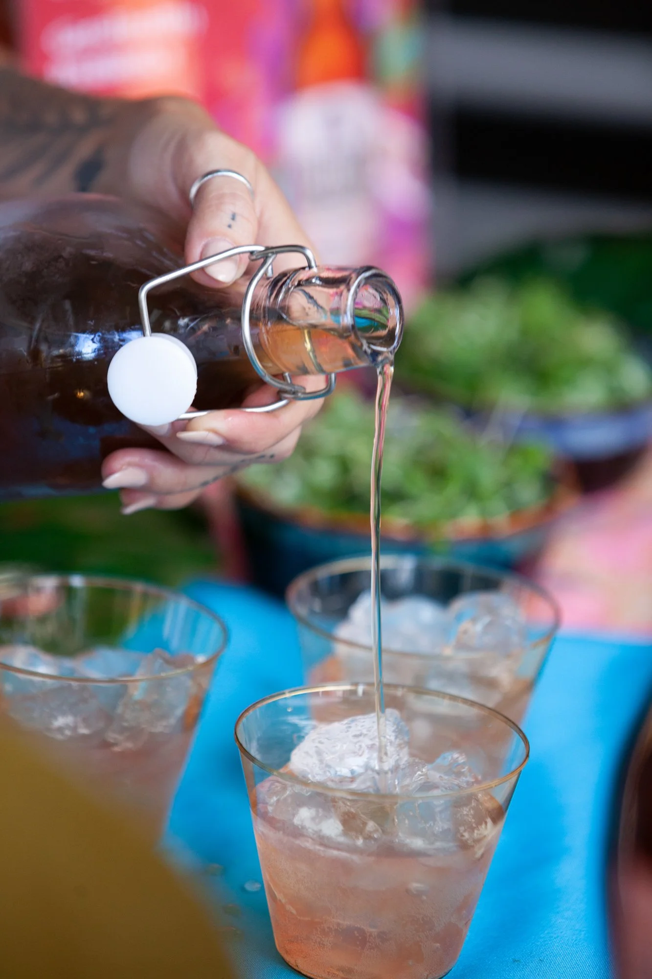 A hand pours pink-colored soda from a glass bottle into a glass filled with ice, with other glasses and bowls of greens in the background.