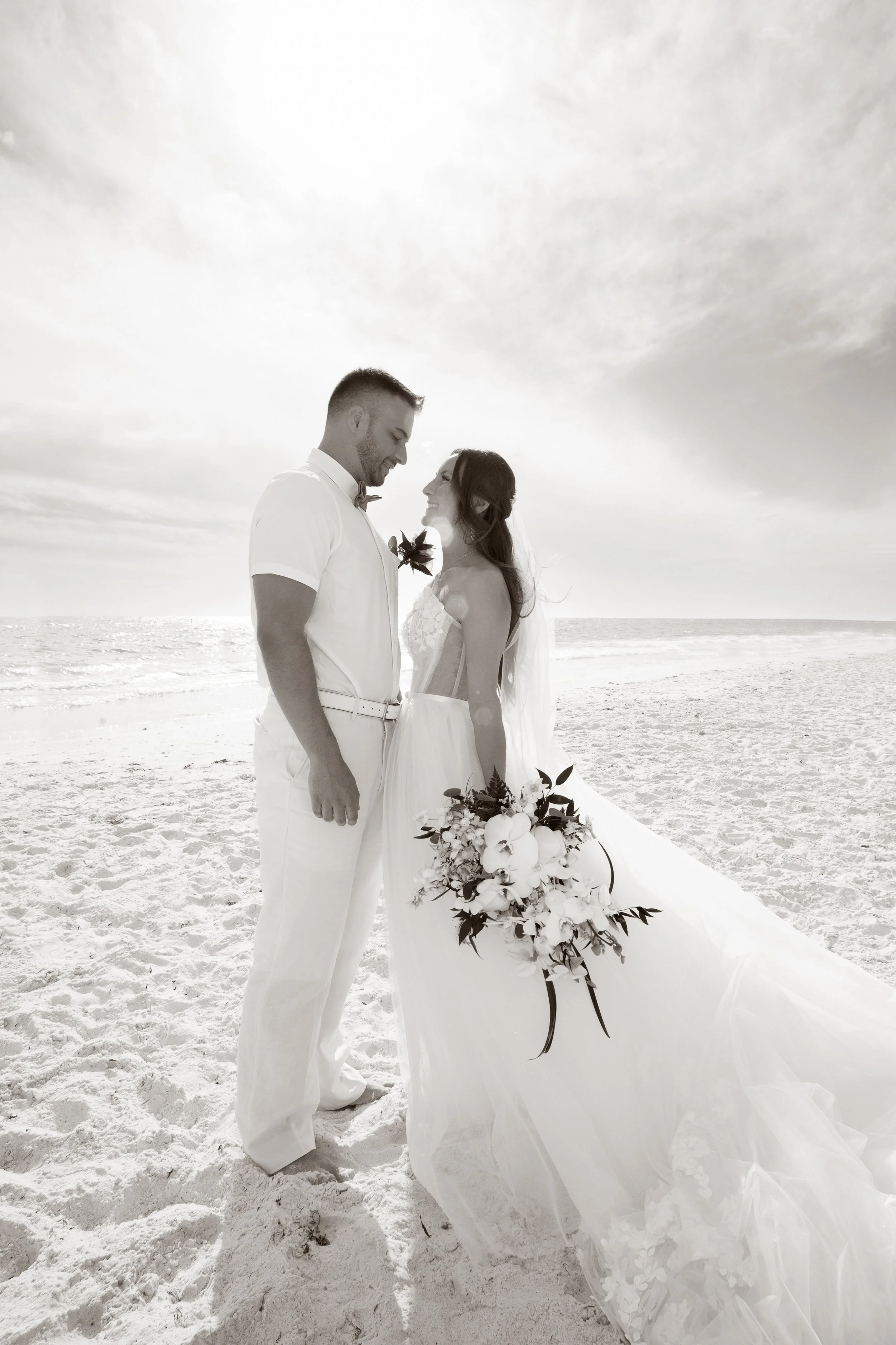 A wedding couple stands on the beach, facing each other and smiling, with the ocean in the background in a black and white photograph.