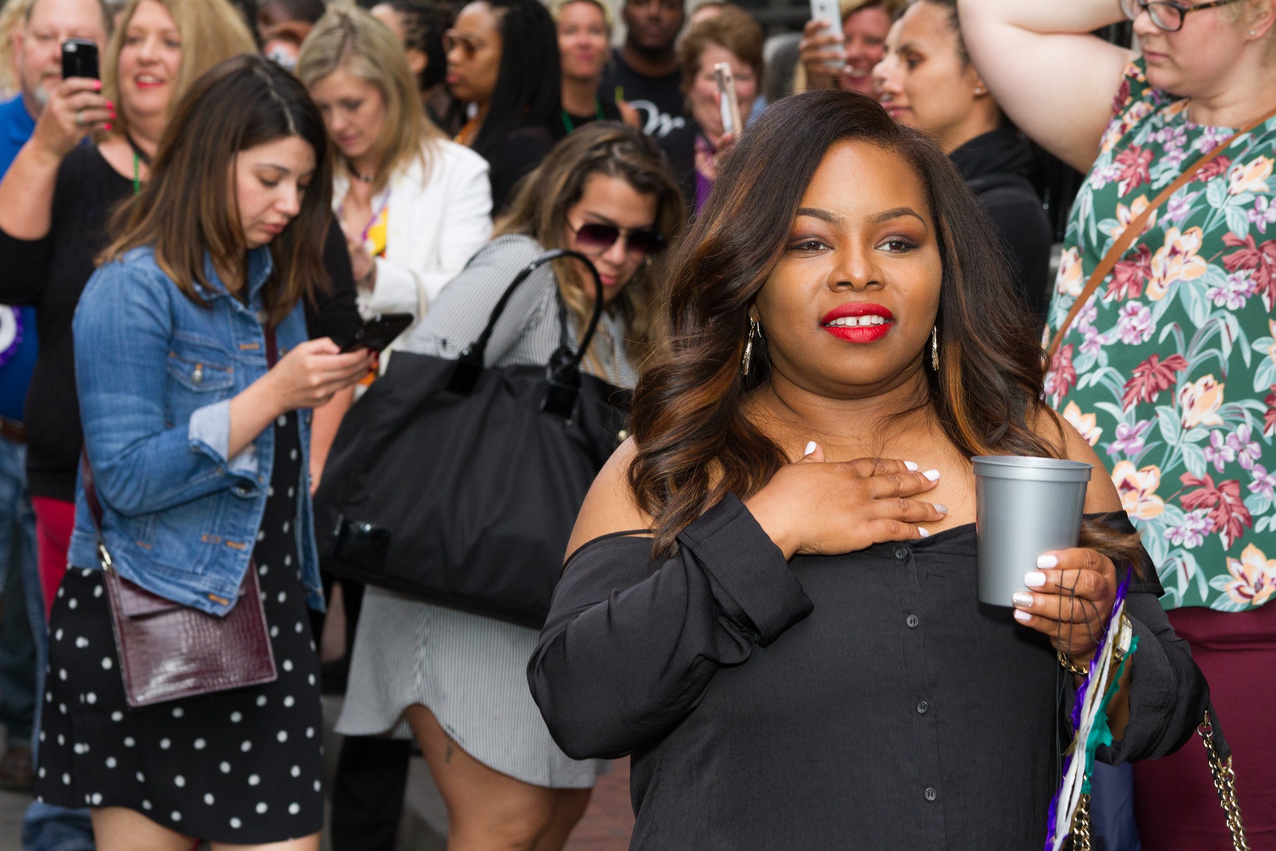 A woman with shoulder-length wavy hair, wearing a black off-the-shoulder top and red lipstick, is holding a gray cup and has her right hand on her chest, with a crowd of people in the background, some using their phones.