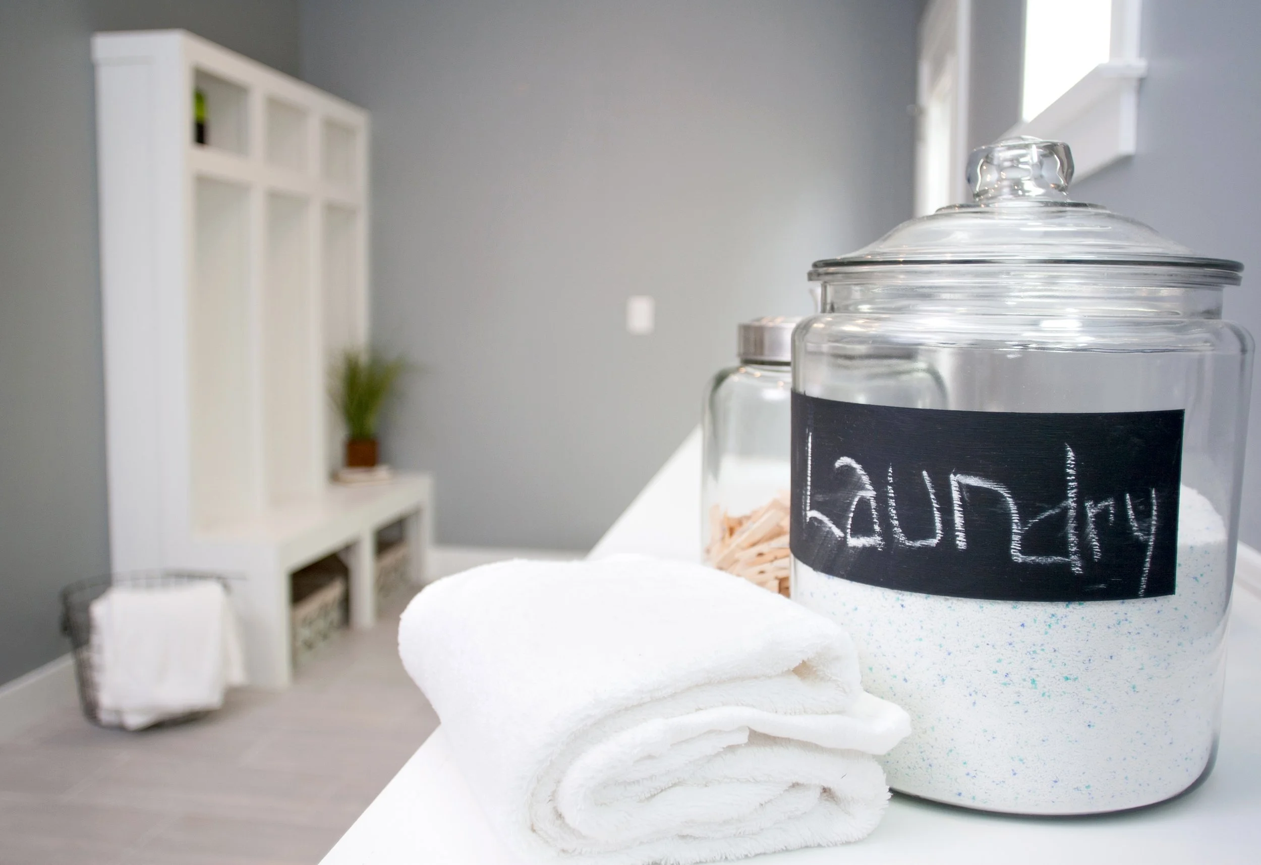 Laundry supplies on a white counter, including a glass jar labeled "Laundry" with white detergent, a smaller jar with sticks, a rolled white towel, and a blurred background of a grey wall, white shelf, and a basket with towels.