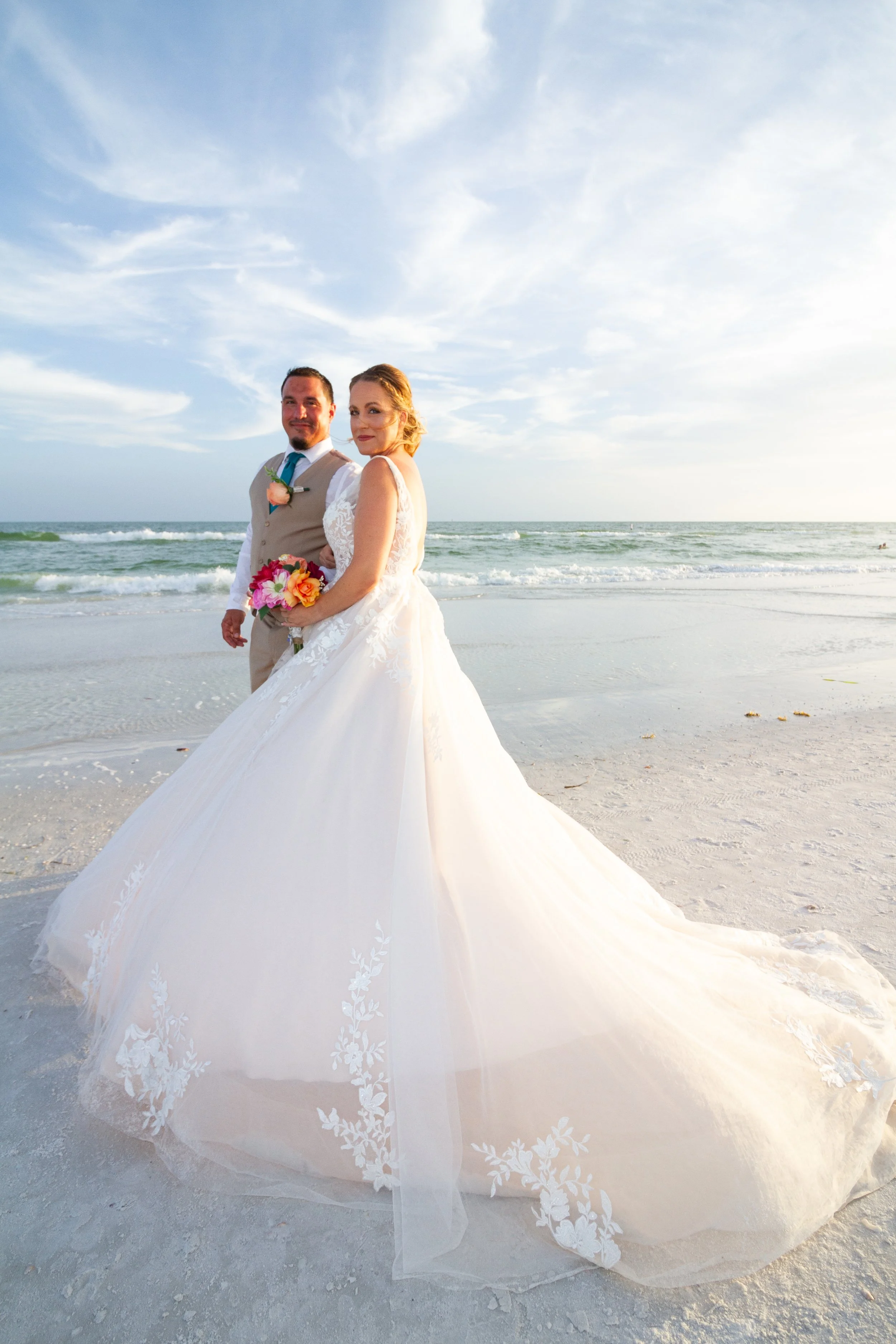 A bride and groom standing on a beach during sunset, dressed in wedding attire with the bride holding a colorful bouquet, behind ocean waves.