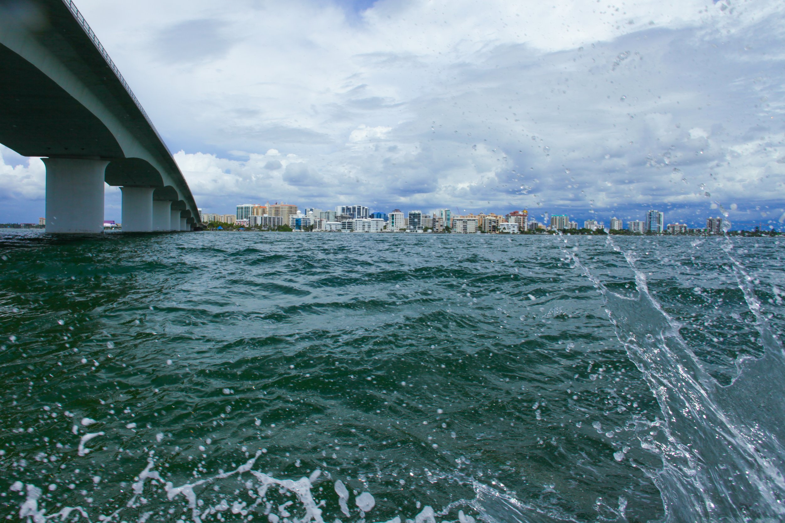 View of a city skyline across water with a bridge on the left, water splashing in the foreground, and a cloudy sky above.