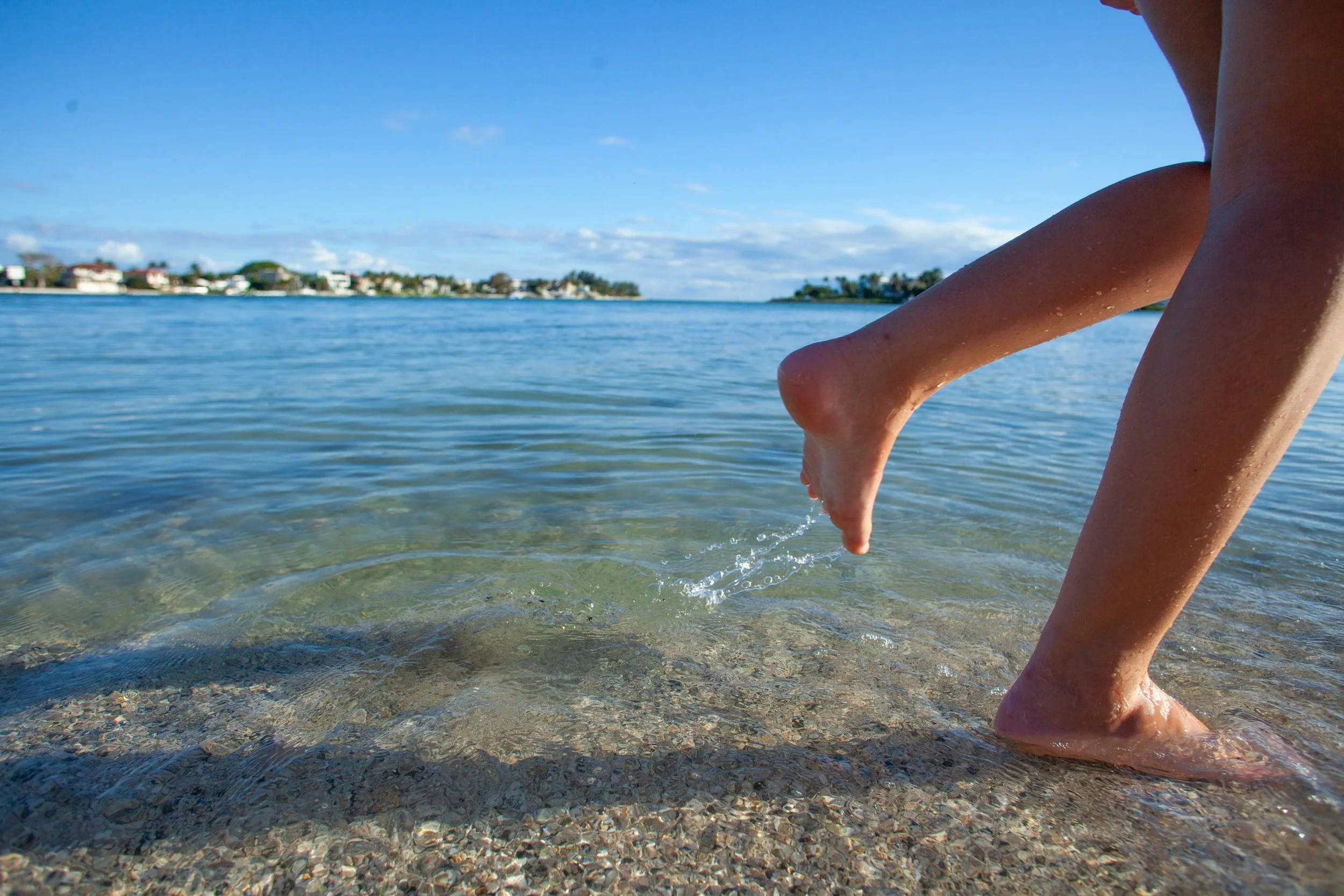 Child's legs and feet playing in shallow water at the beach with houses in the background