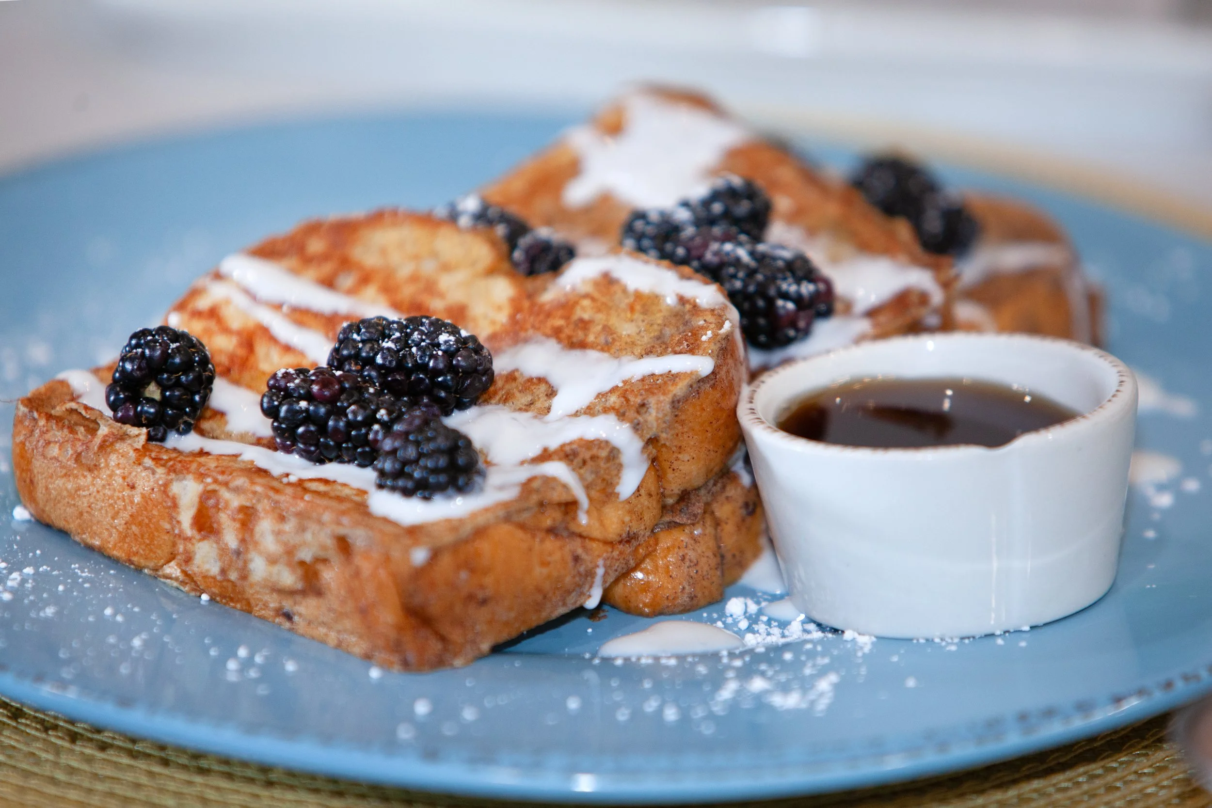 French toast topped with blackberries and drizzled with syrup, served with a small cup of syrup on a blue plate.