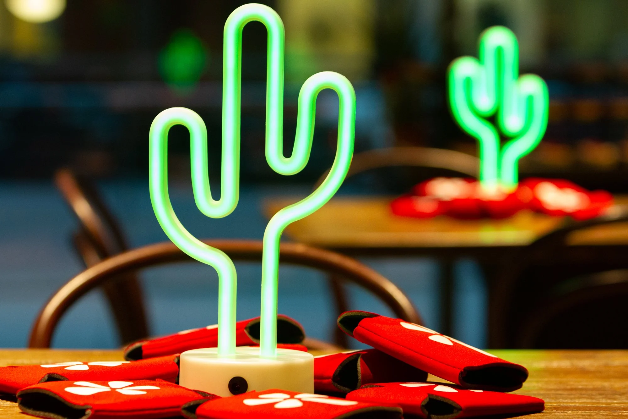 Neon cactus-shaped light on a wooden table surrounded by red napkins, with another neon cactus in the background.