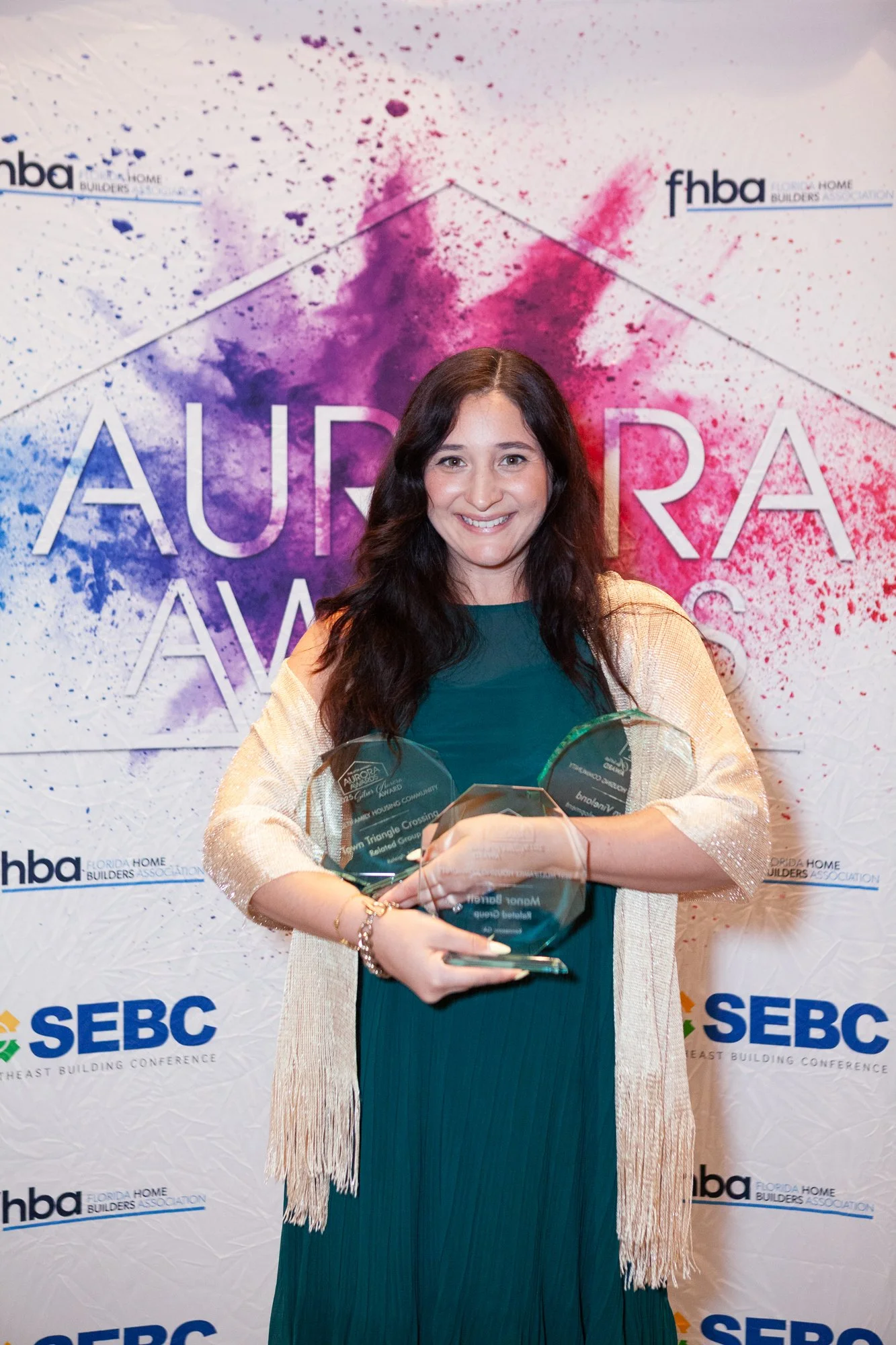 A woman with dark wavy hair smiling and holding multiple awards in front of a backdrop with logos and colorful paint splashes. She is wearing a teal dress and a beige fringed shawl.