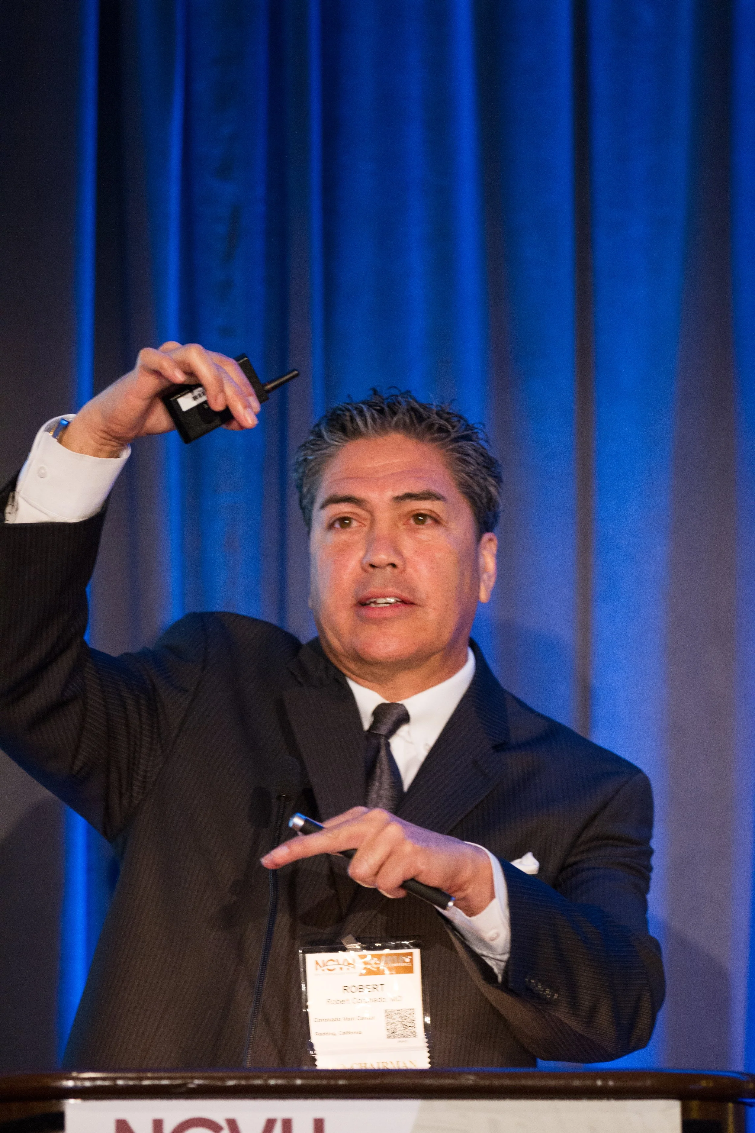 A man in a dark suit and tie speaking at a conference, holding a microphone in one hand and a remote in the other, with a blue curtain background.