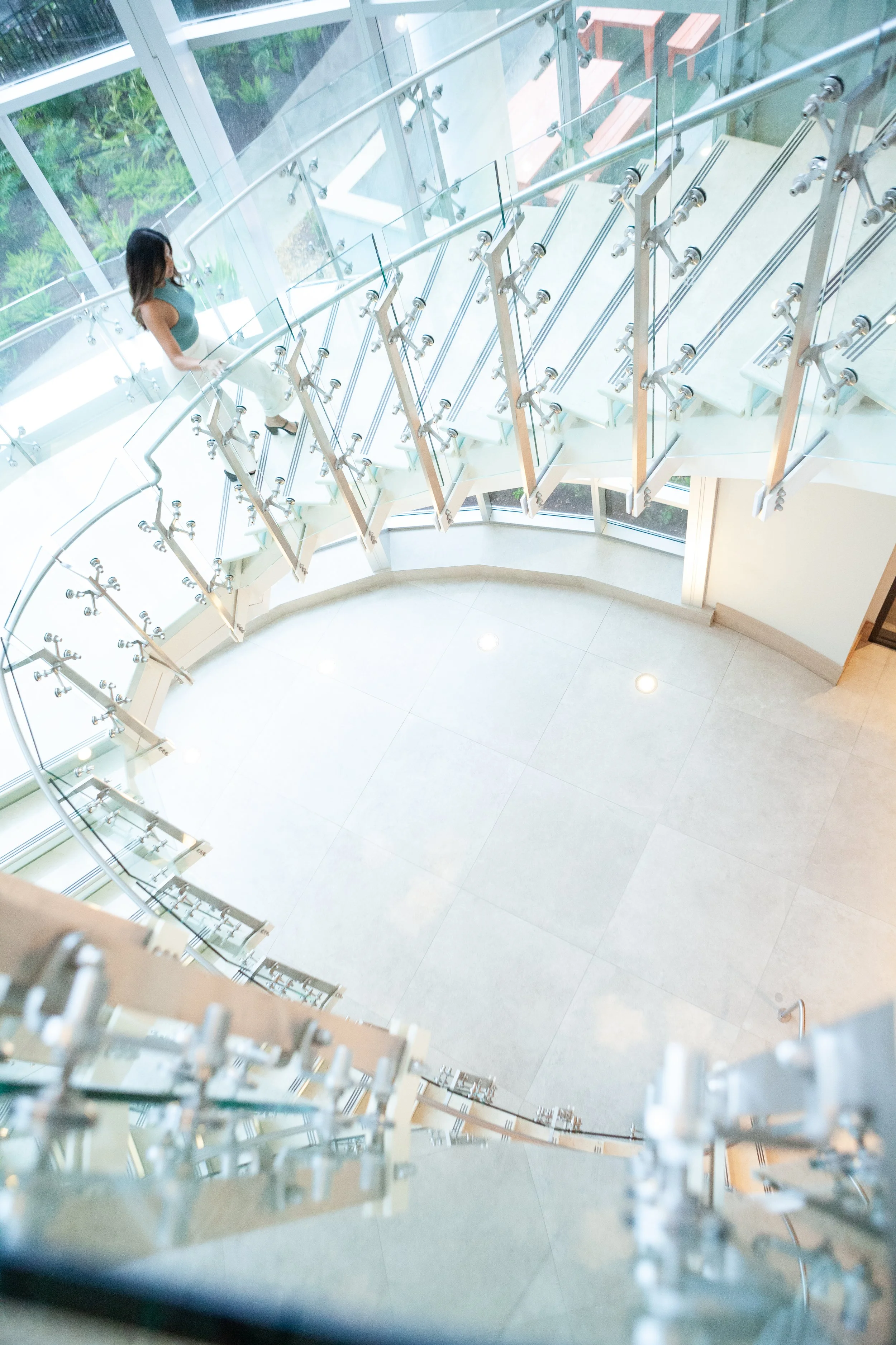 Inside view of a modern building with a staircase, glass railings, and large windows revealing greenery outside. A woman is walking on the stairs.