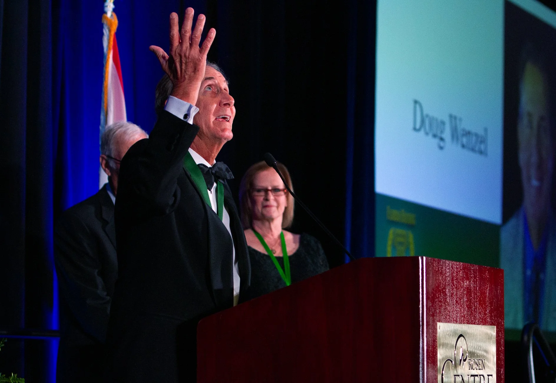 An older man in a black tuxedo and bow tie is speaking at a podium during a formal event. He is raising his right hand, and a woman with glasses and a black dress stands behind him. A man with glasses is partially visible behind him. The background h