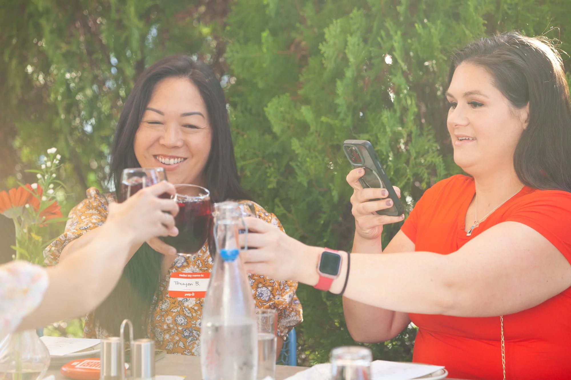 Two women clinking glasses of red wine at an outdoor gathering, one is taking a selfie or photo with her phone, on a sunny day with greenery in the background.