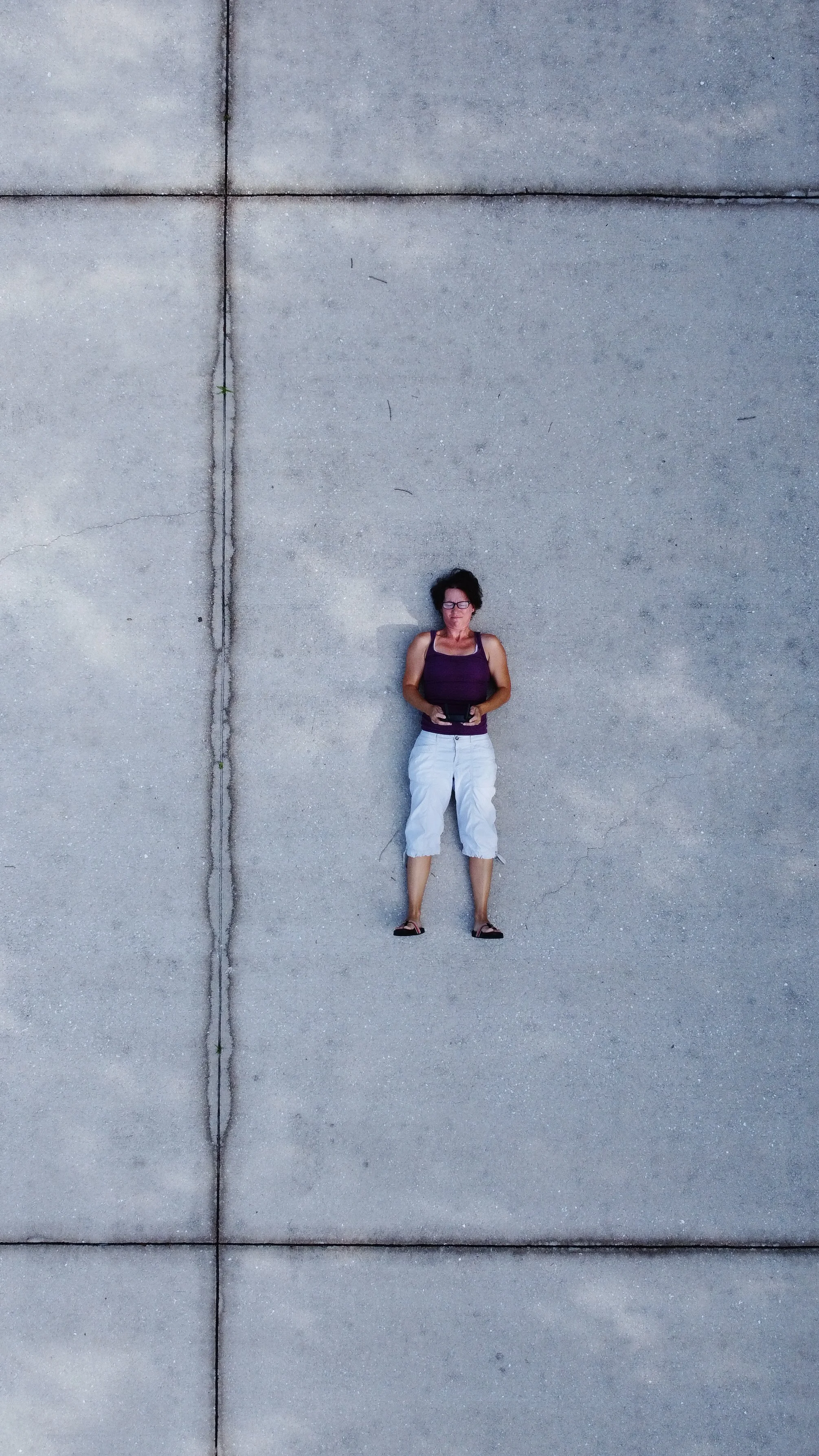 Woman lying on concrete sidewalk, looking up, wearing glasses, a purple tank top, white capri pants, and black sandals, holding a tablet in her hands.