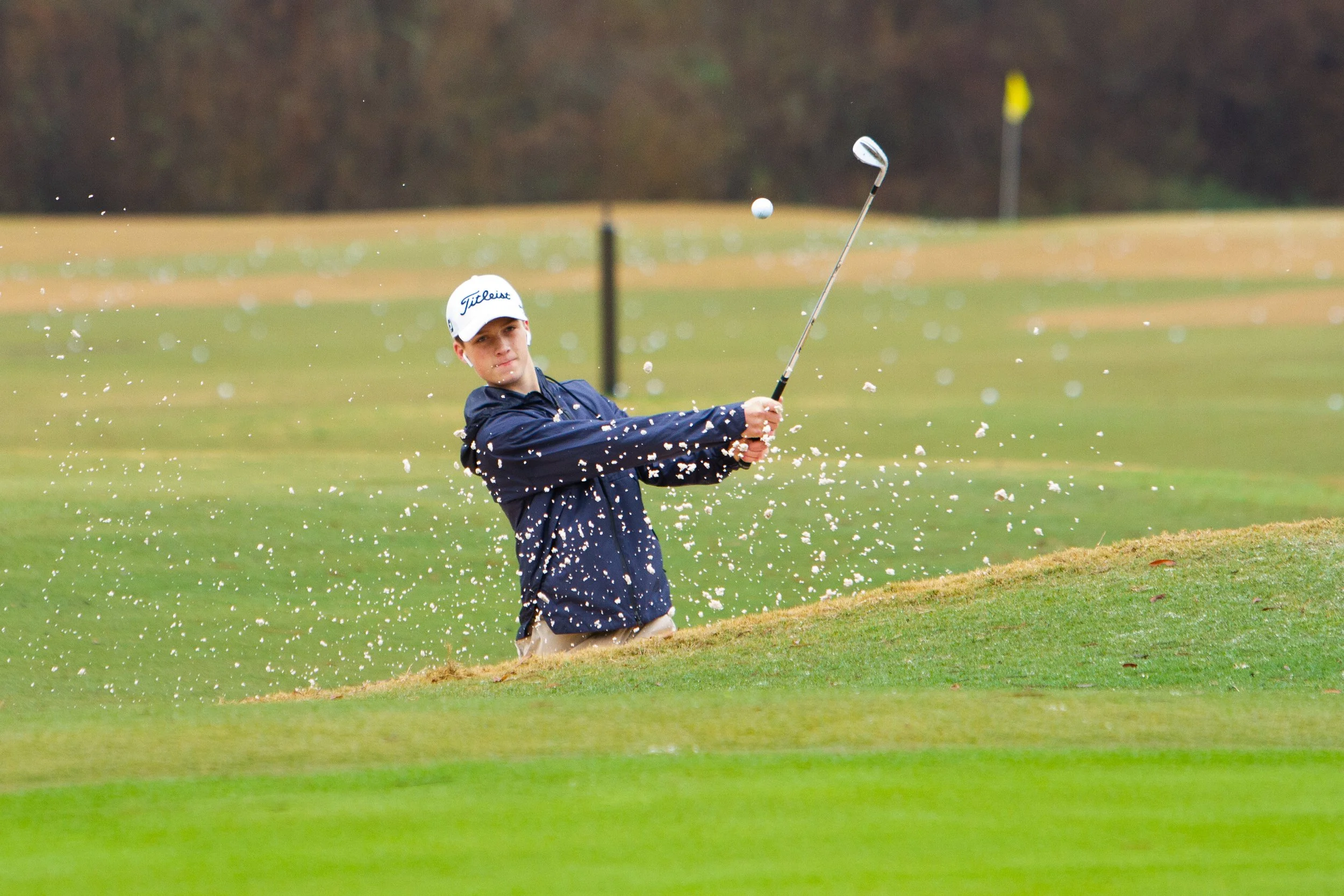 A young man in a navy jacket and white cap is hitting a golf ball from a sand bunker on a golf course, with sand flying around him.