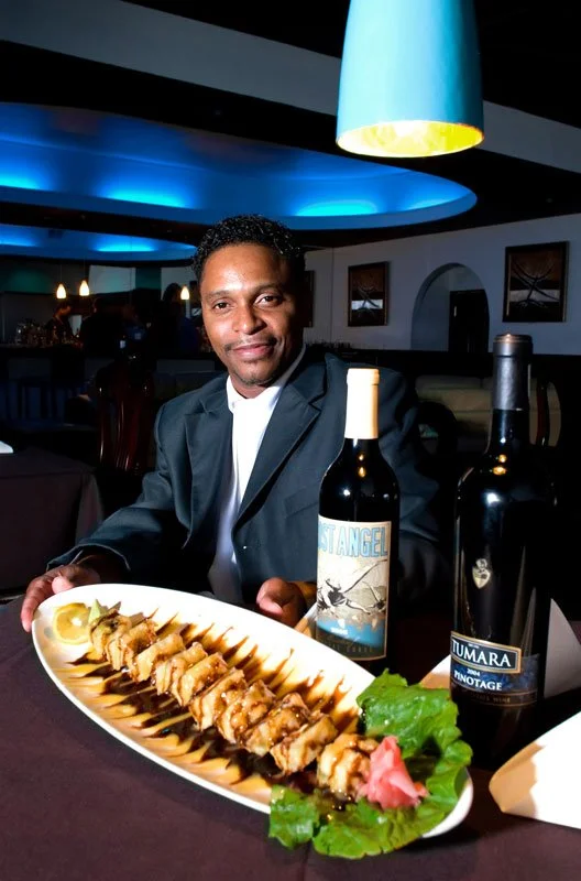 A man in a suit dining at a restaurant, with a plate of sushi, a bottle of wine labeled 'St. Angel,' and a bottle of Lumara wine on the table.