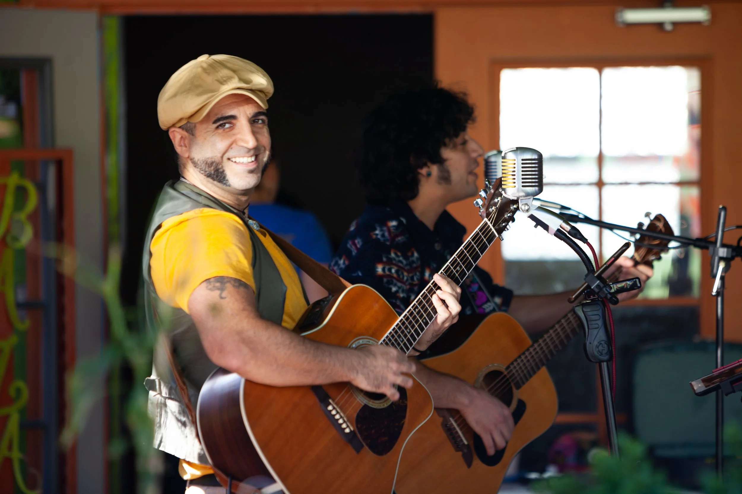 Two musicians playing acoustic guitars in a cozy indoor setting, with one smiling directly at the camera.