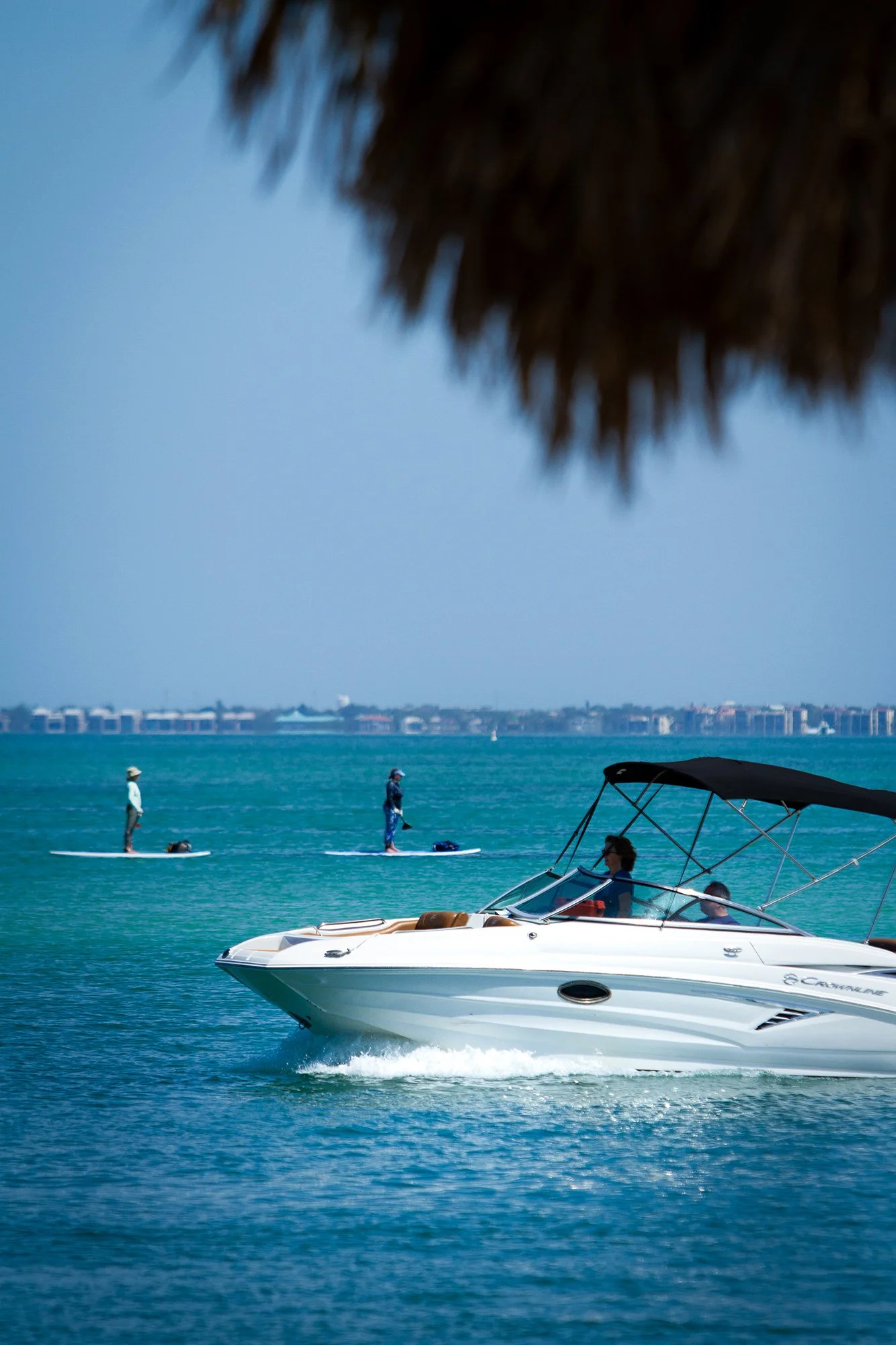 A white motorboat on turquoise water, with a person onboard, passing near three paddleboarders in the distance, under a blue sky.