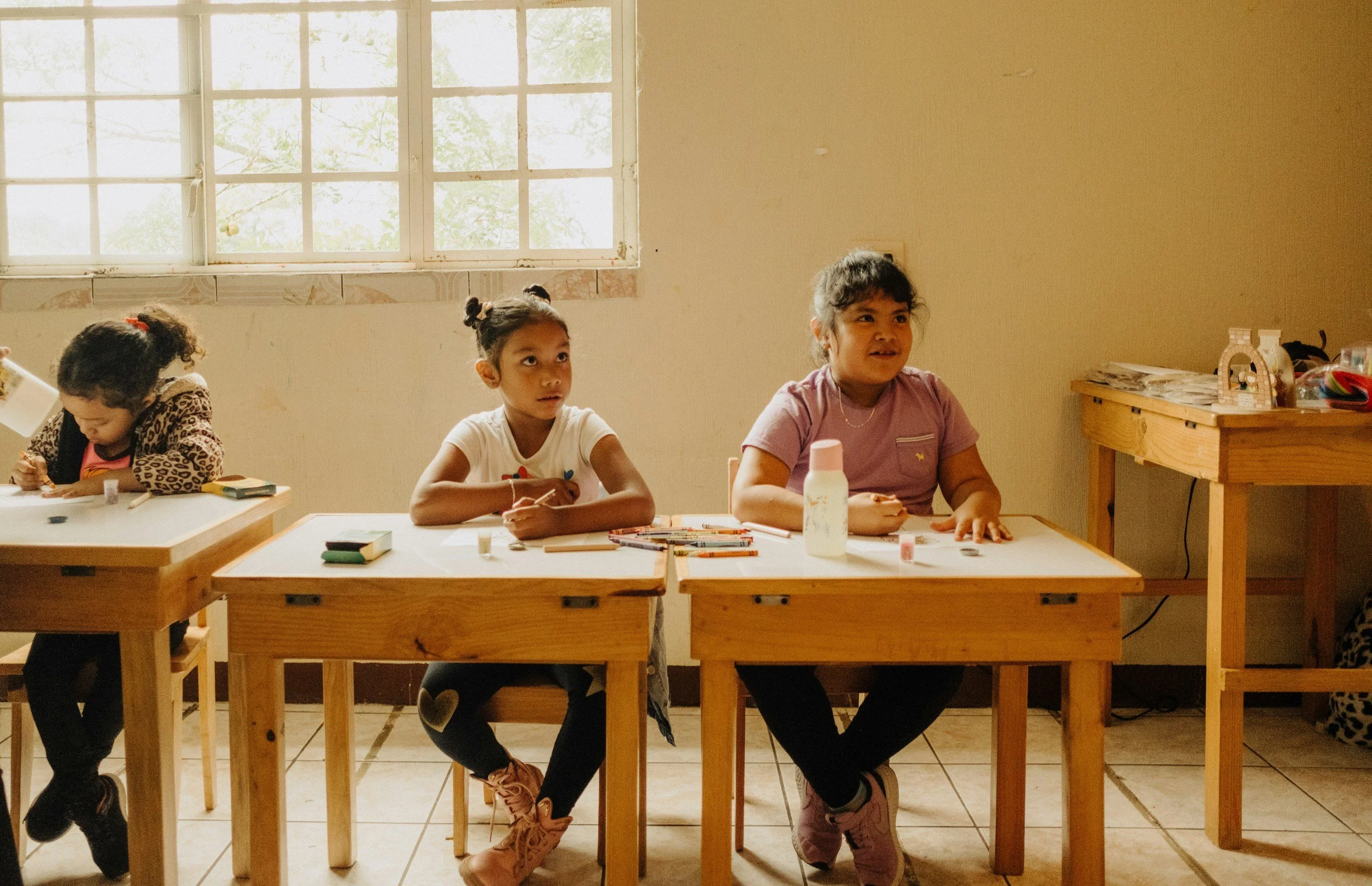 Three young girls sitting at wooden desks in a classroom with a window behind them. The girls are engaged in activities, with various small objects and books on their desks. The classroom has tiled floor and a plain wall.