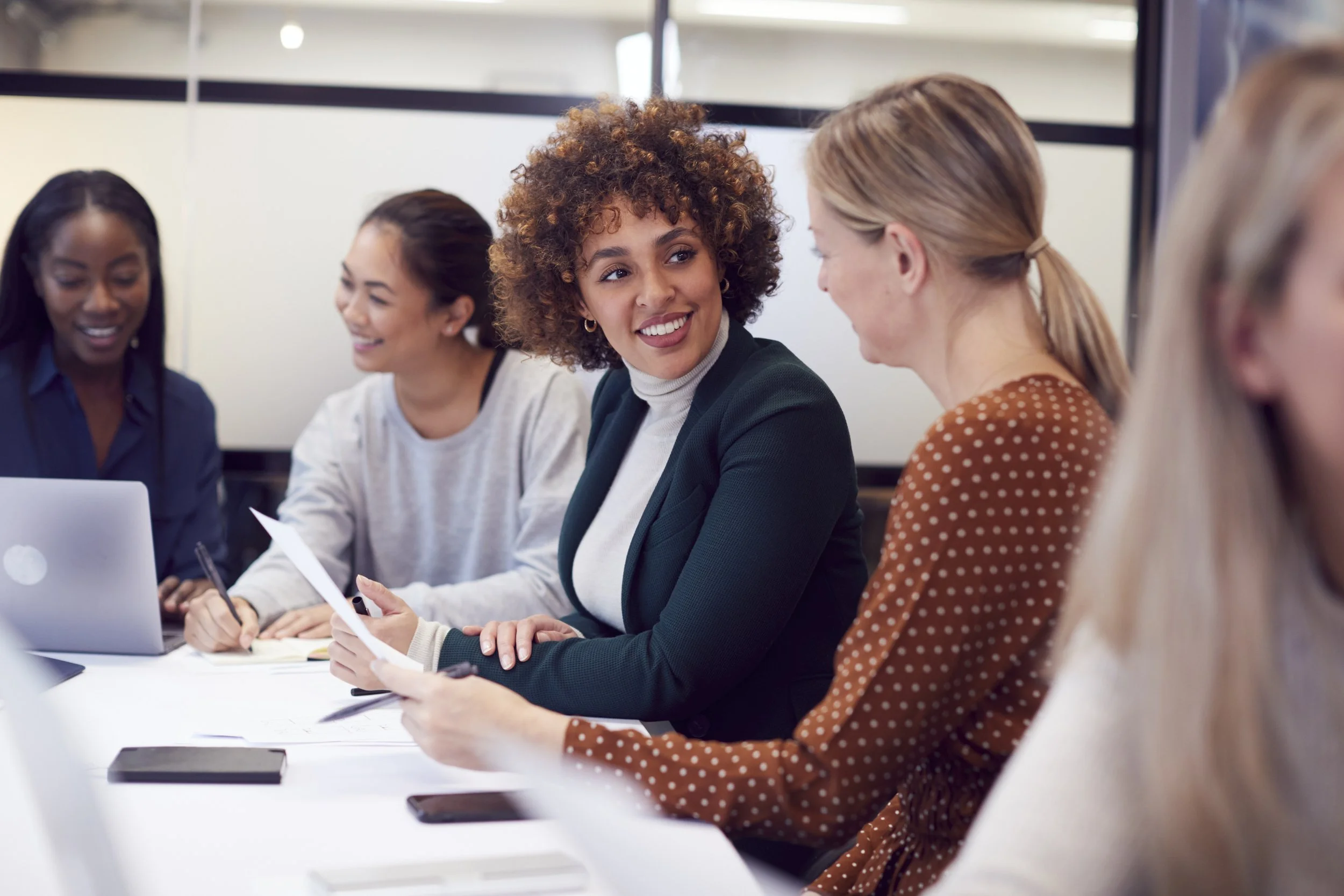 A diverse group of women are sitting around a table, engaged in a discussion in a modern office setting. They are smiling and appear to be having a productive meeting.