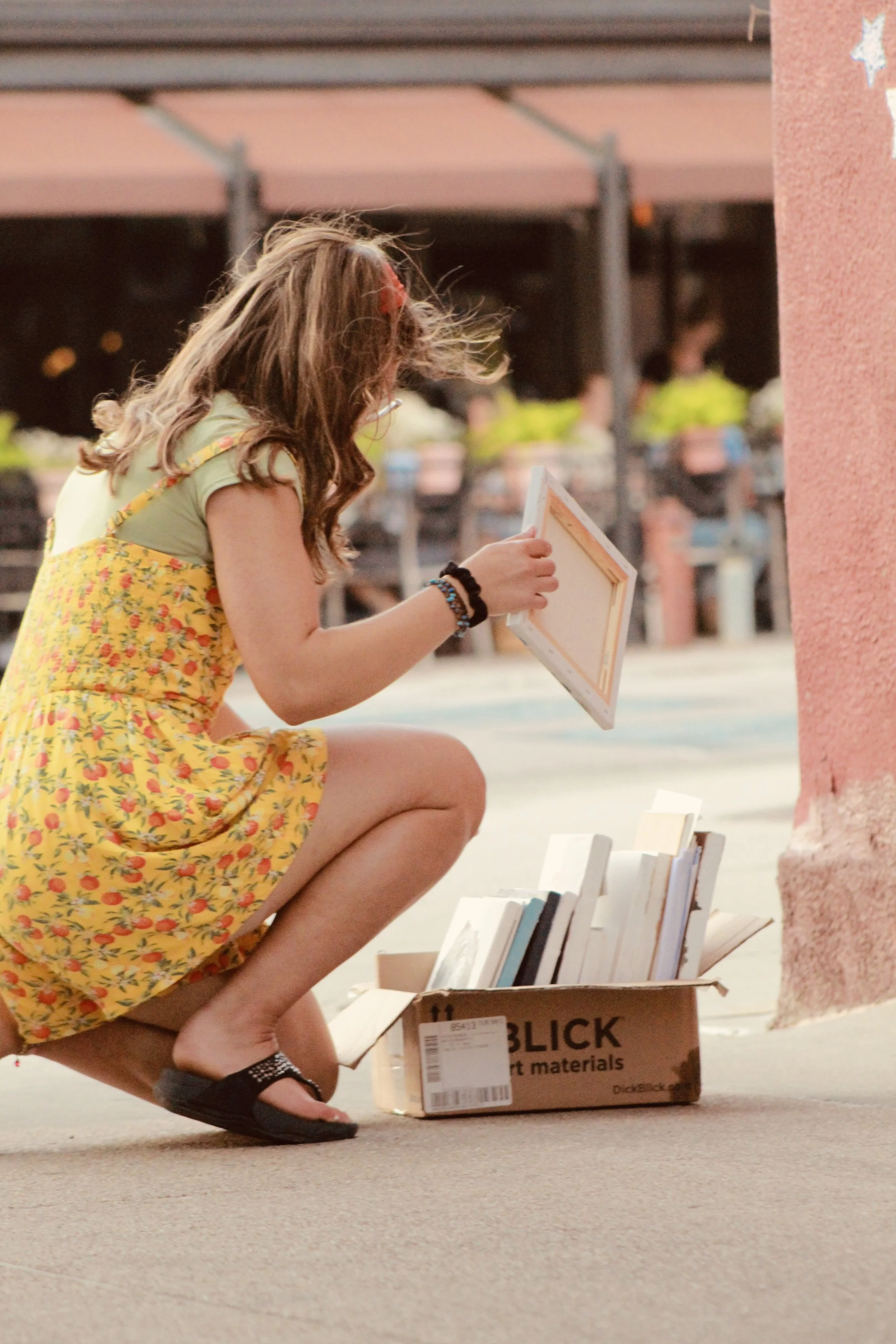 A young girl with long brown hair, wearing a yellow floral dress and black sandals, is crouching down and looking at books displayed on the ground. She appears to be reading or examining the books, which are in an open cardboard box marked 'BLICK art materials.' The setting is indoors, with a person in a yellow shirt and other people in the background.