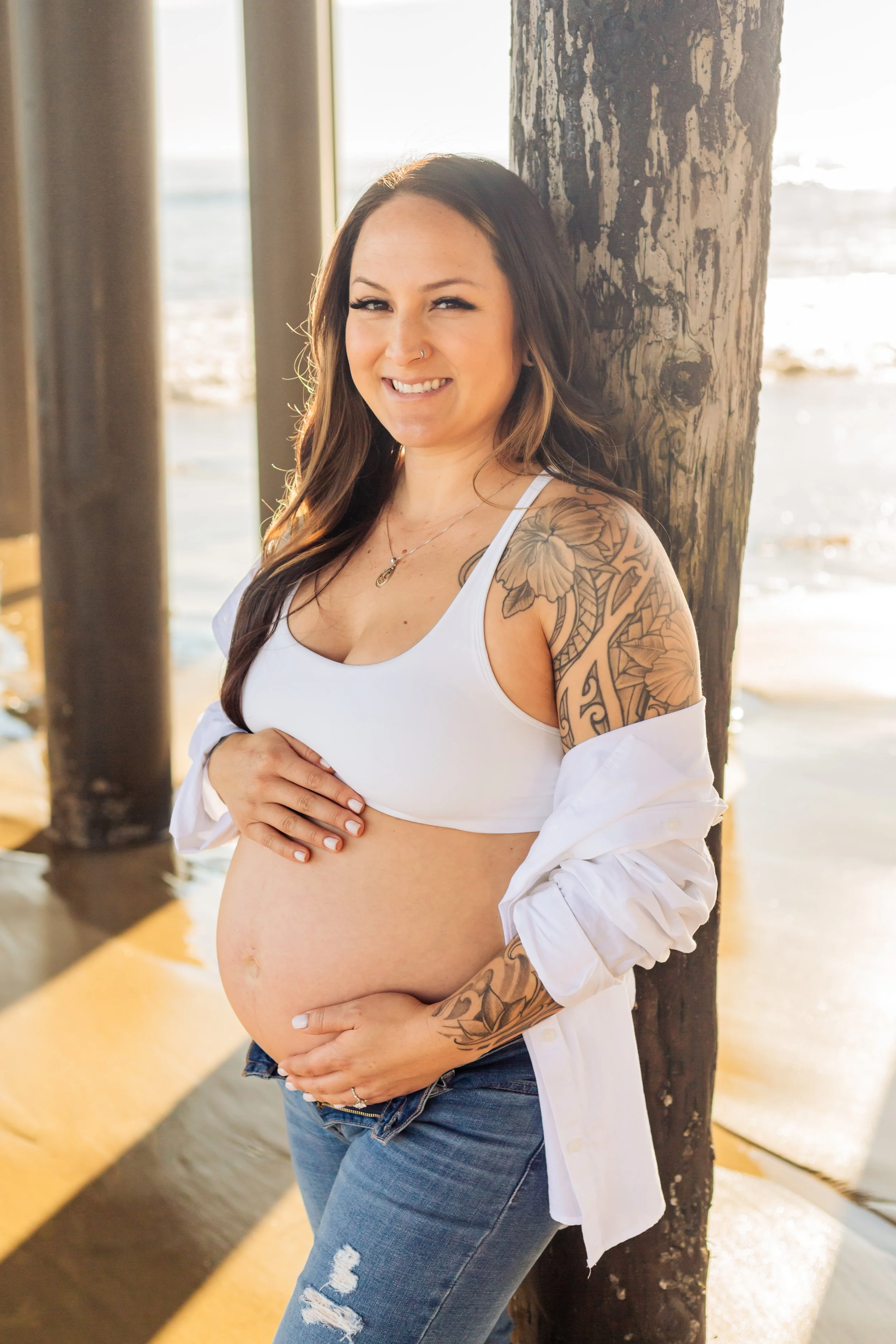 A pregnant woman with tattoos on her arm and shoulder, wearing a white crop top and jeans, standing near a wooden post on a beach. She is smiling, with her hand on her belly, with the ocean and sunset in the background.