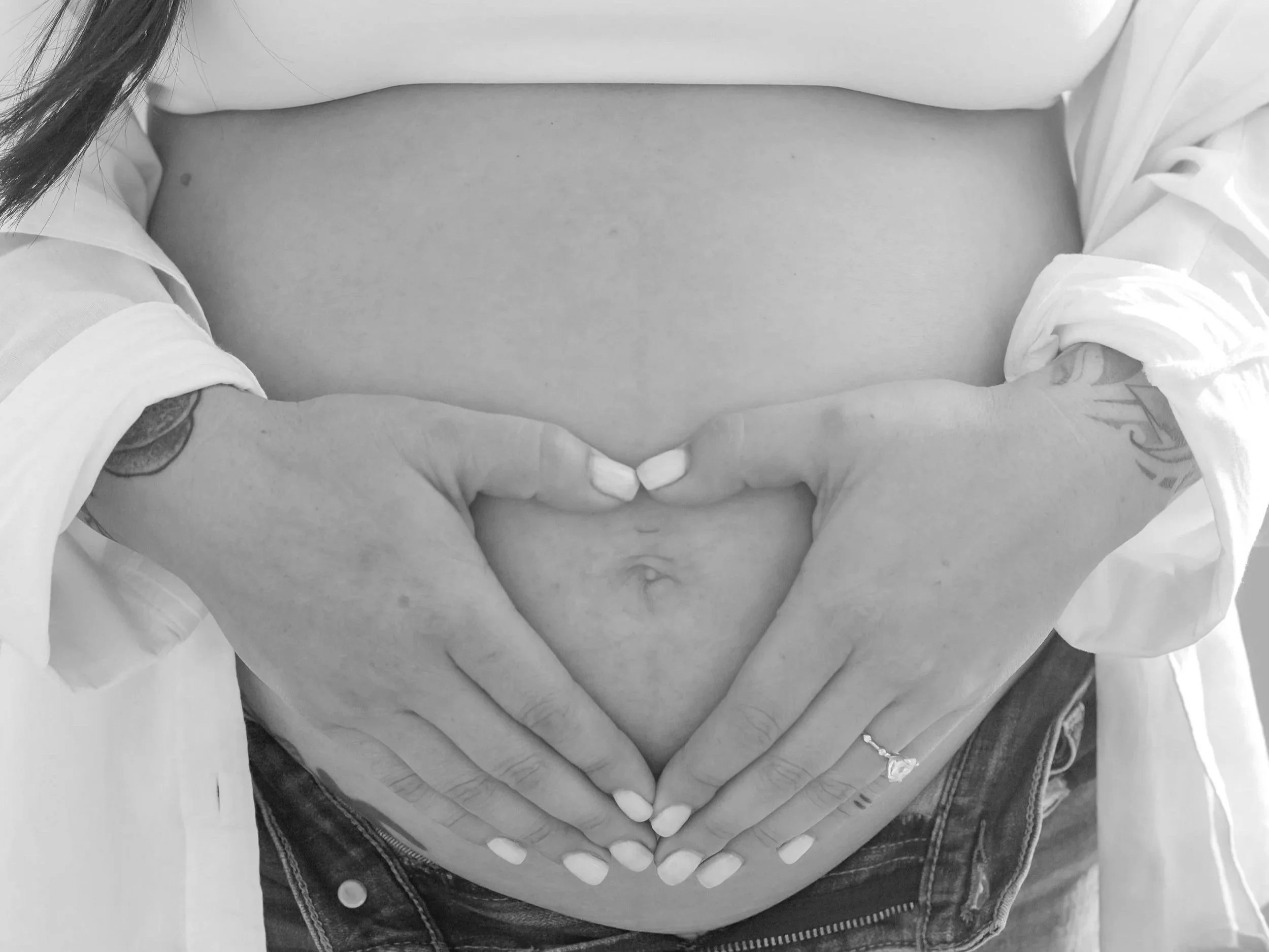 Black and white photo of a pregnant woman with her hands in the shape of a heart on her belly, wearing a white shirt and denim jeans.