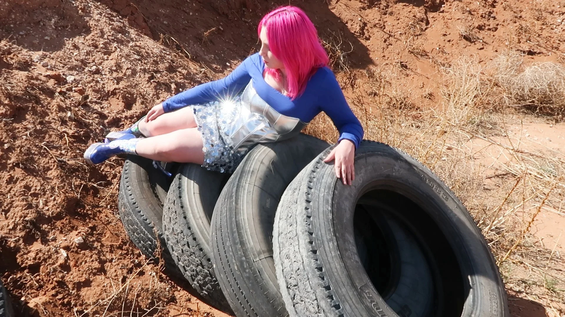 Woman with pink hair wearing a blue top, silver skirt, and blue shoes sitting on stacked tires in a desert area with reddish soil and dry plants.