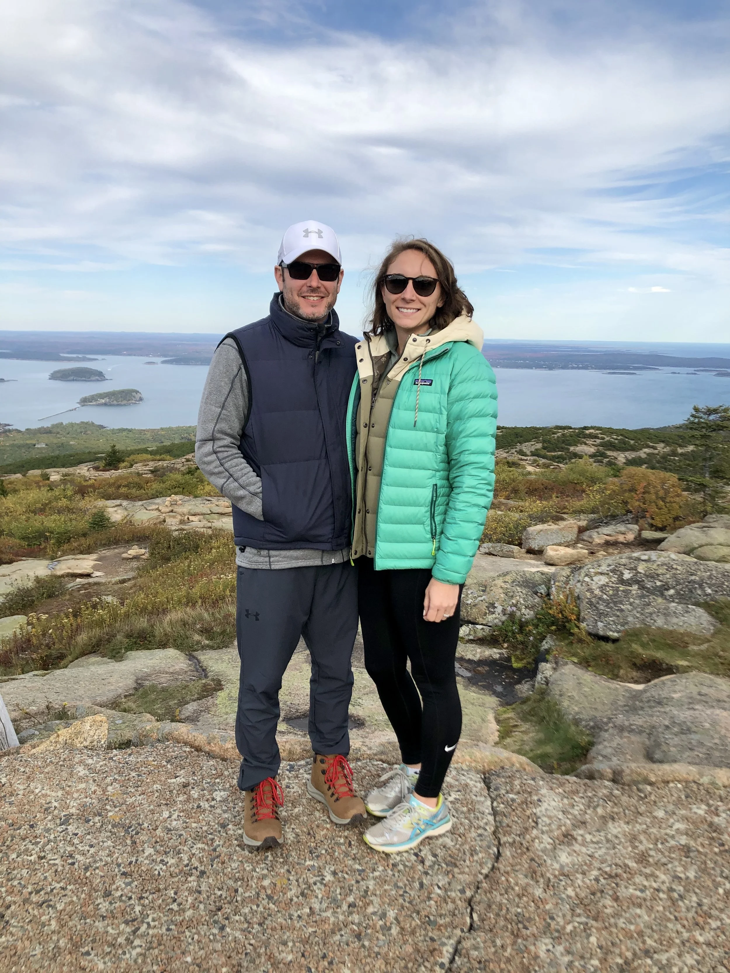 A smiling man and woman standing on a rocky hilltop with a scenic view of a large body of water and small islands in the background, dressed in outdoor clothing and sunglasses.