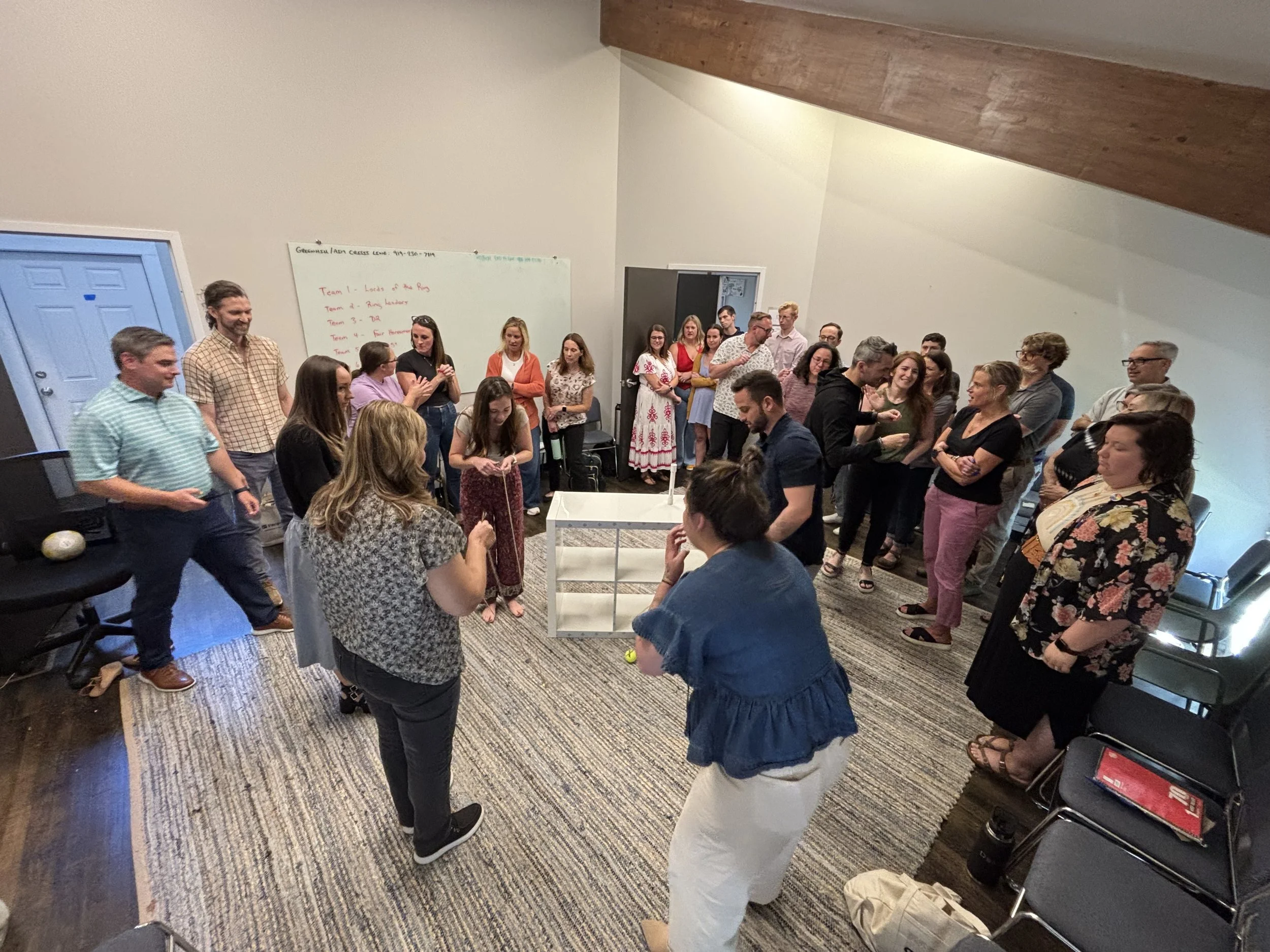 Group of people standing and sitting in a meeting room with a whiteboard in the background. Some individuals are engaged in conversation while others are observing.