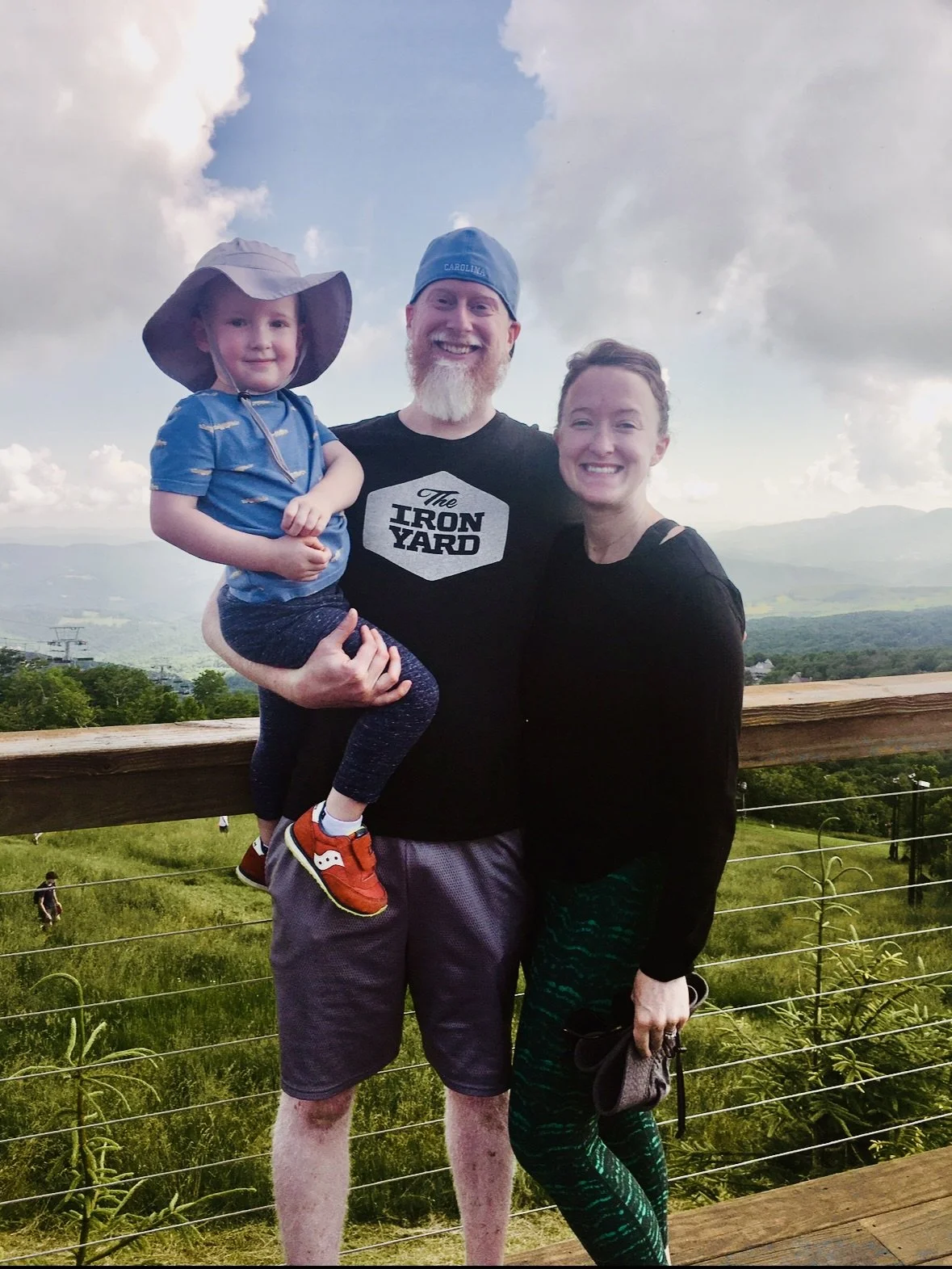 Three people, a man, a woman, and a young child, standing on a wooden deck with a mountainous landscape behind them. The man is smiling and holding the child, who is wearing a sun hat and bright orange shoes. The woman is standing next to them, smiling. The sky is partly cloudy.