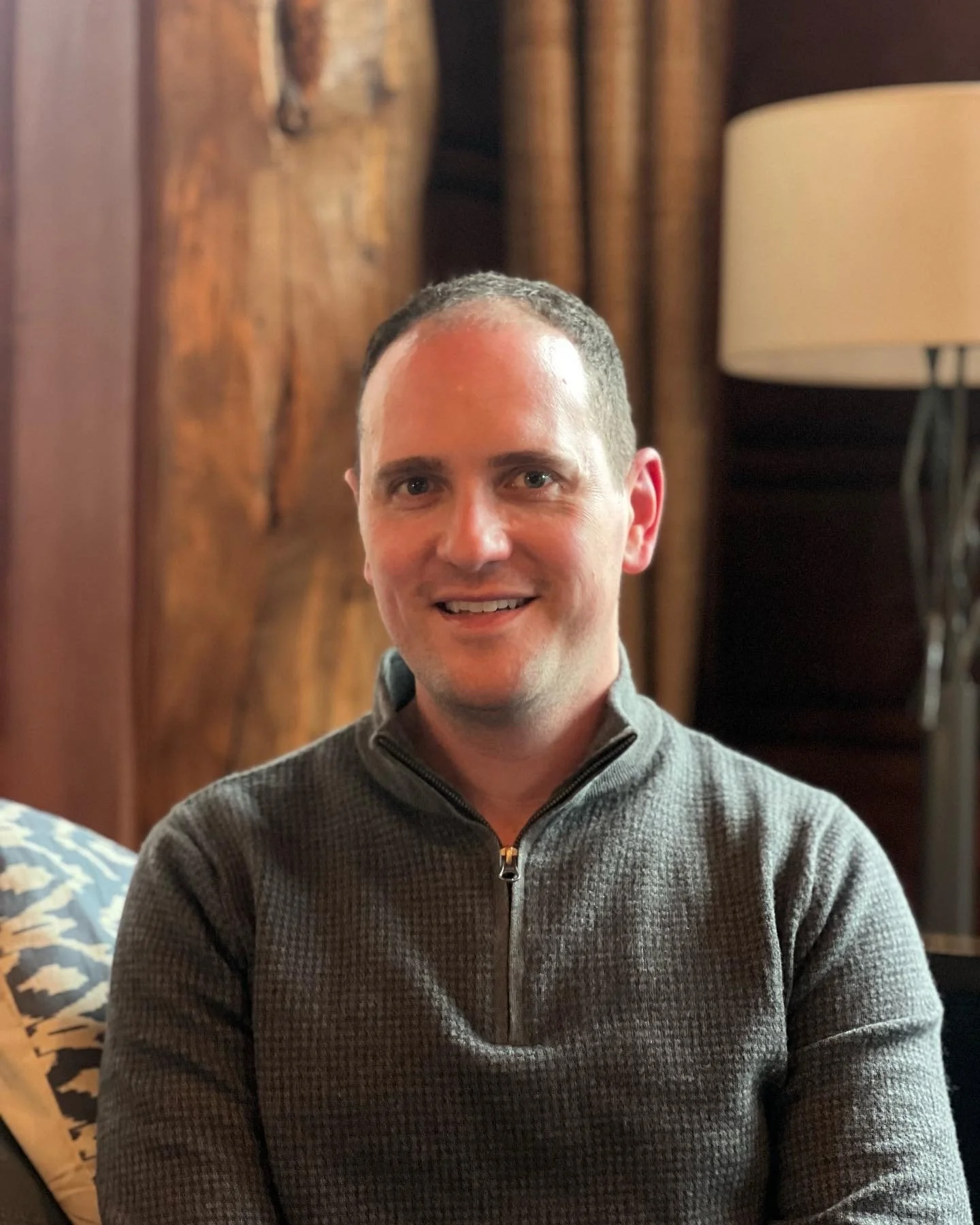 A smiling man with short dark hair and light skin, wearing a gray zip-up sweater, sitting indoors with wooden wall panels and a beige lamp in the background.