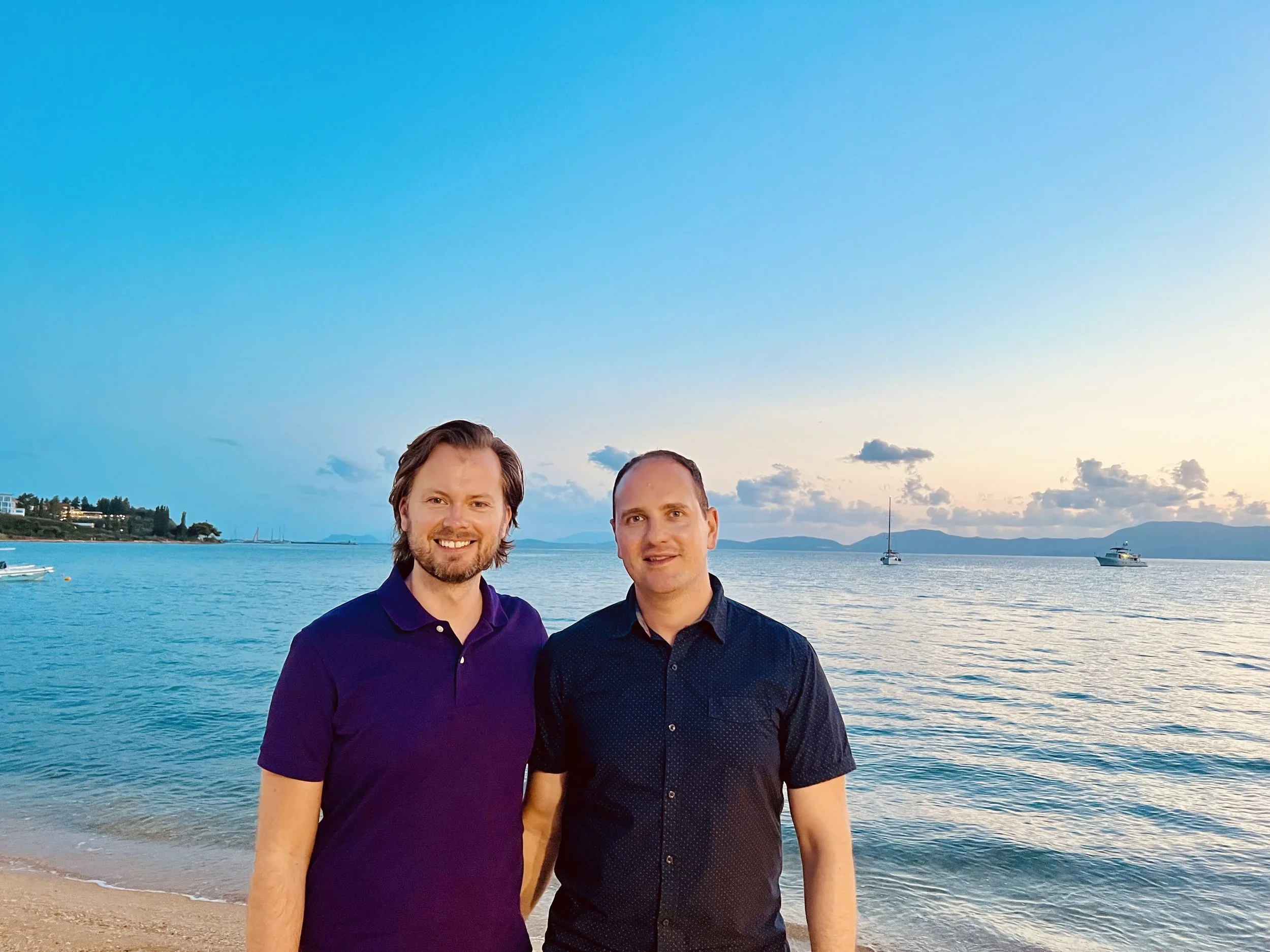 Two men standing on a beach at sunset with water and sailboats in the background.