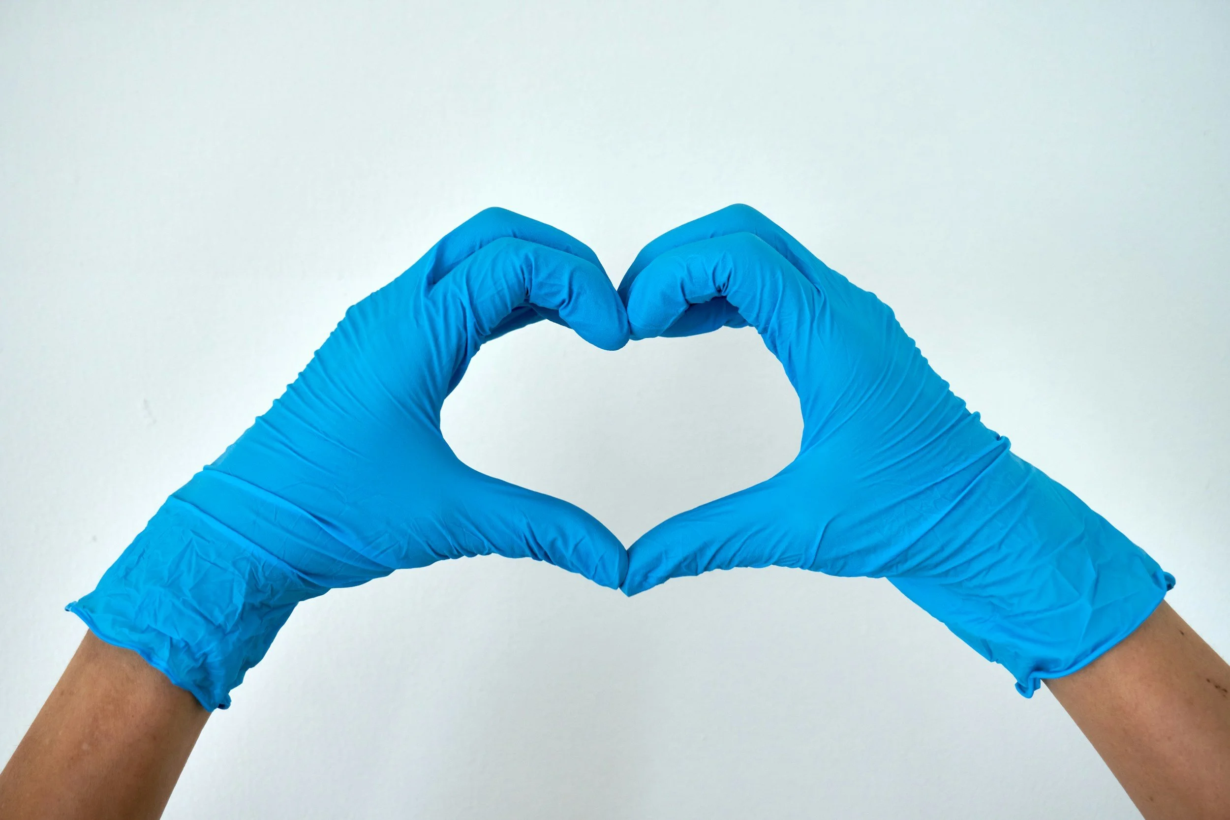 Hands wearing blue gloves forming a heart shape against a plain light background.