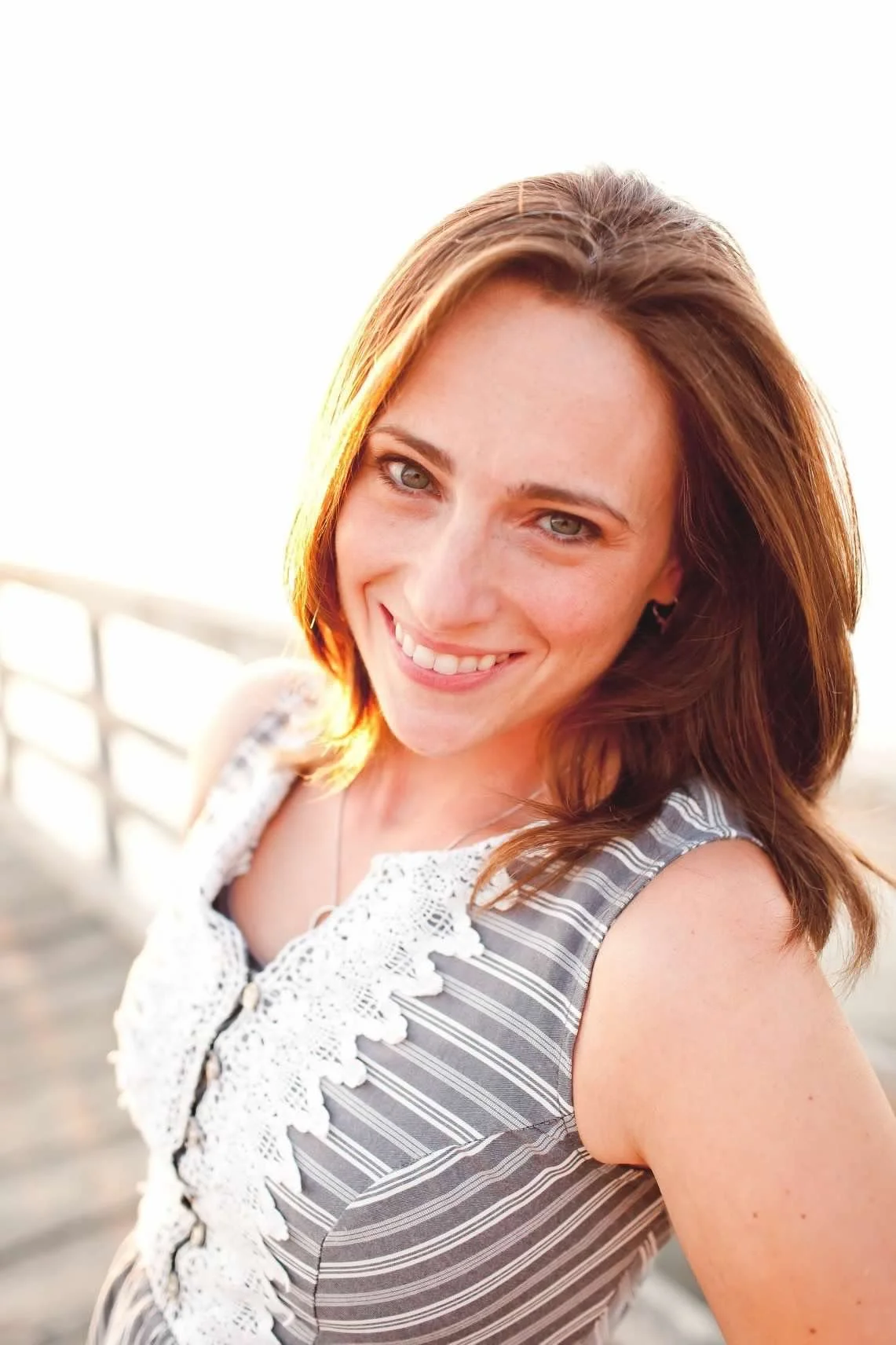 A woman with shoulder-length reddish-brown hair smiling outdoors at sunset, wearing a sleeveless striped top with lace details.